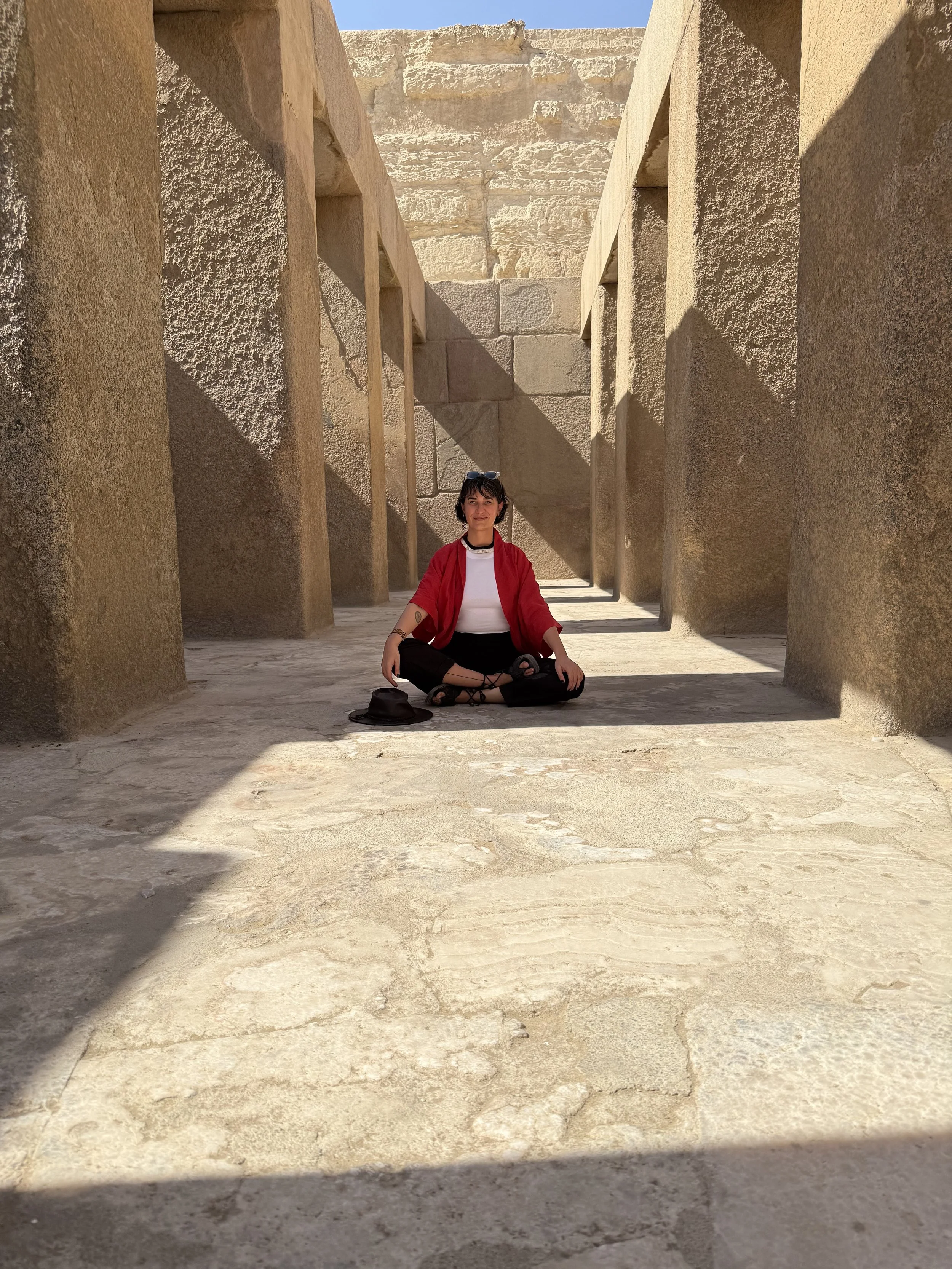 A woman sitting cross-legged on the ground in an ancient stone structure with tall walls and columns, wearing a red jacket, black pants, and sunglasses, with a hat and bag on the ground beside her.