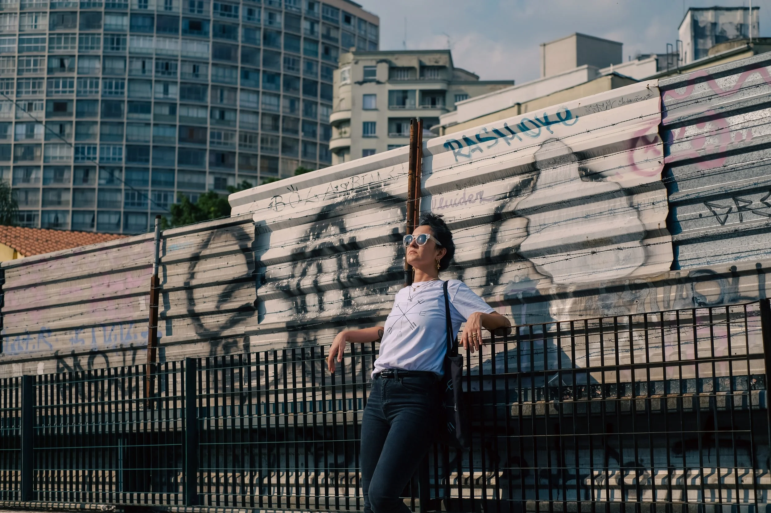 A woman wearing sunglasses standing in front of graffiti-covered metal barricades in an urban area with high-rise buildings in the background.