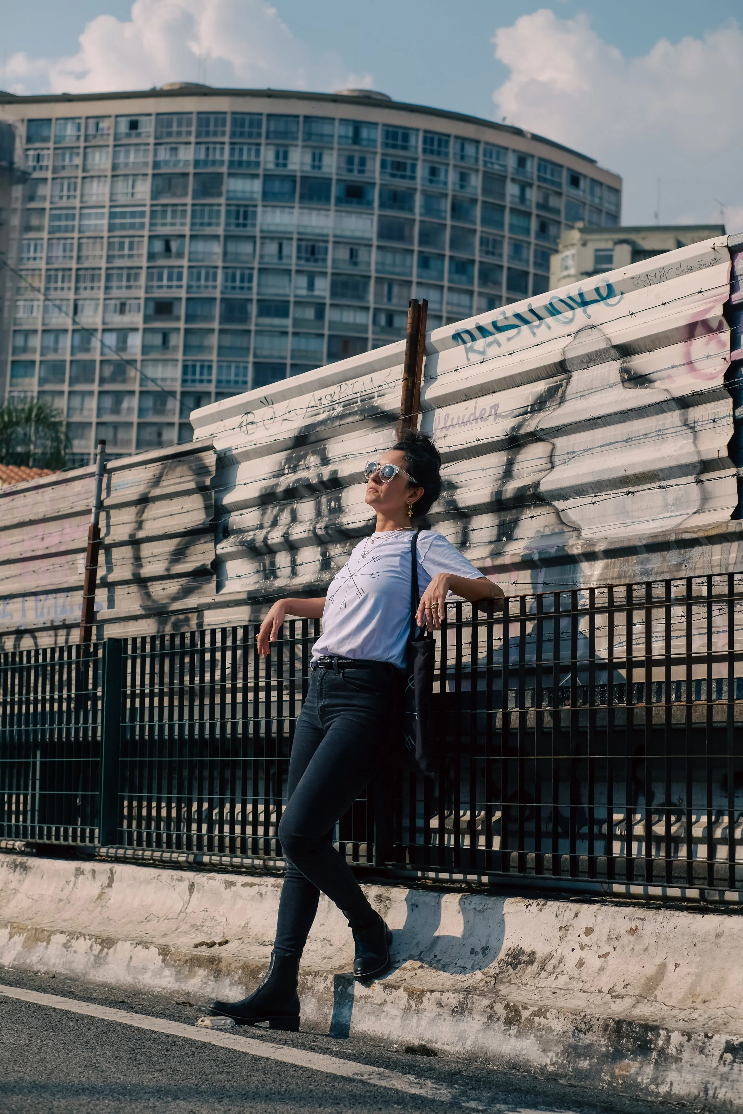 A woman leaning against a fence on a city street with a graffiti-decorated metal sheet behind her and a tall building in the background.