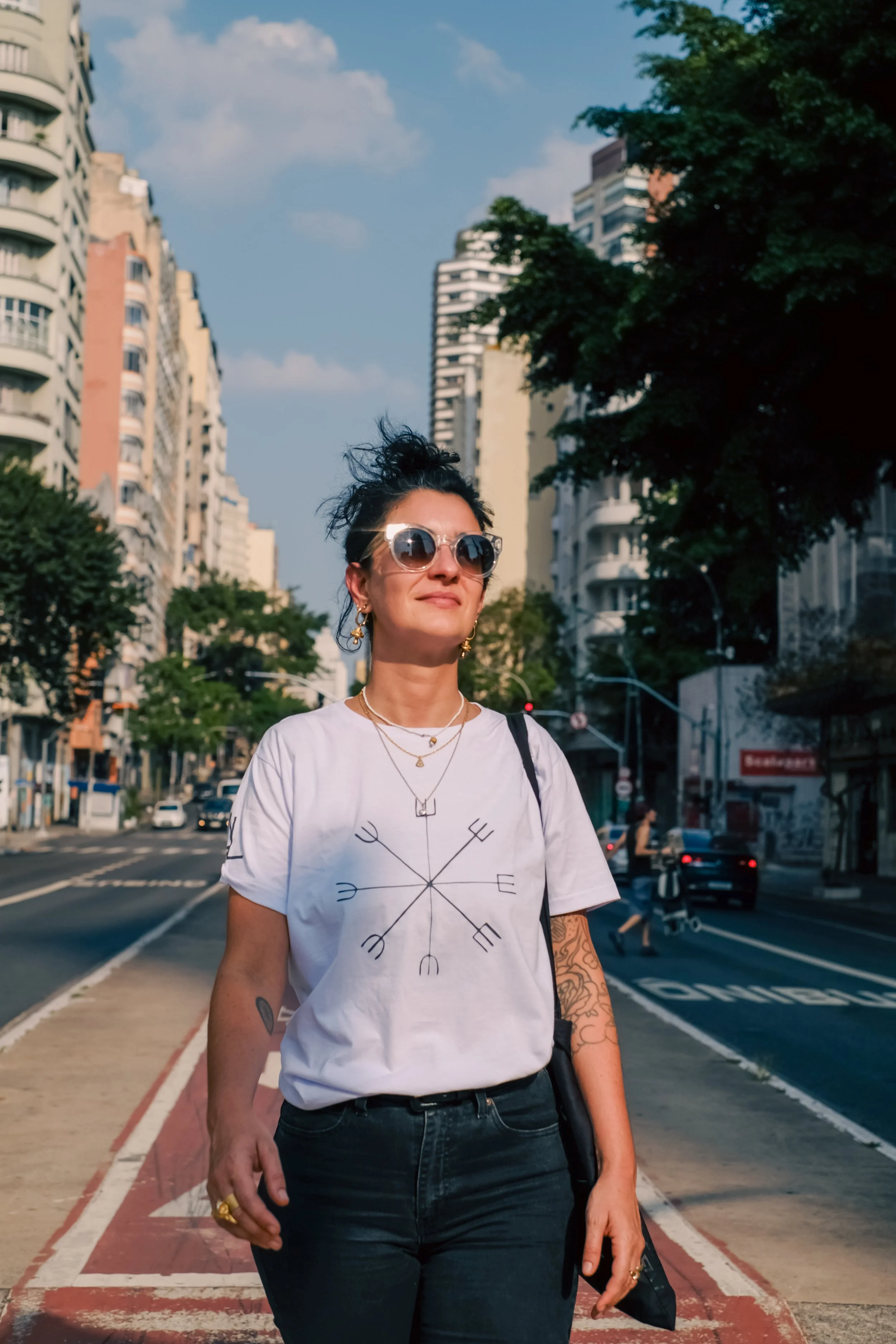 Me wearing sunglasses, a white graphic t-shirt, and black pants stands on a city sidewalk with high-rise buildings and trees in the background.