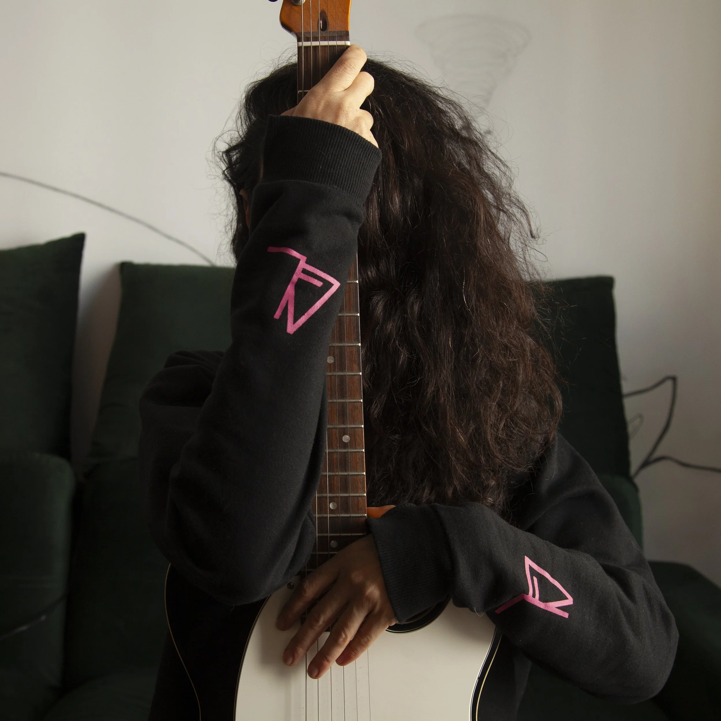 Person with curly hair holding an acoustic guitar, wearing a black sweatshirt with pink logo on sleeves, sitting on a green couch in a room with white walls.