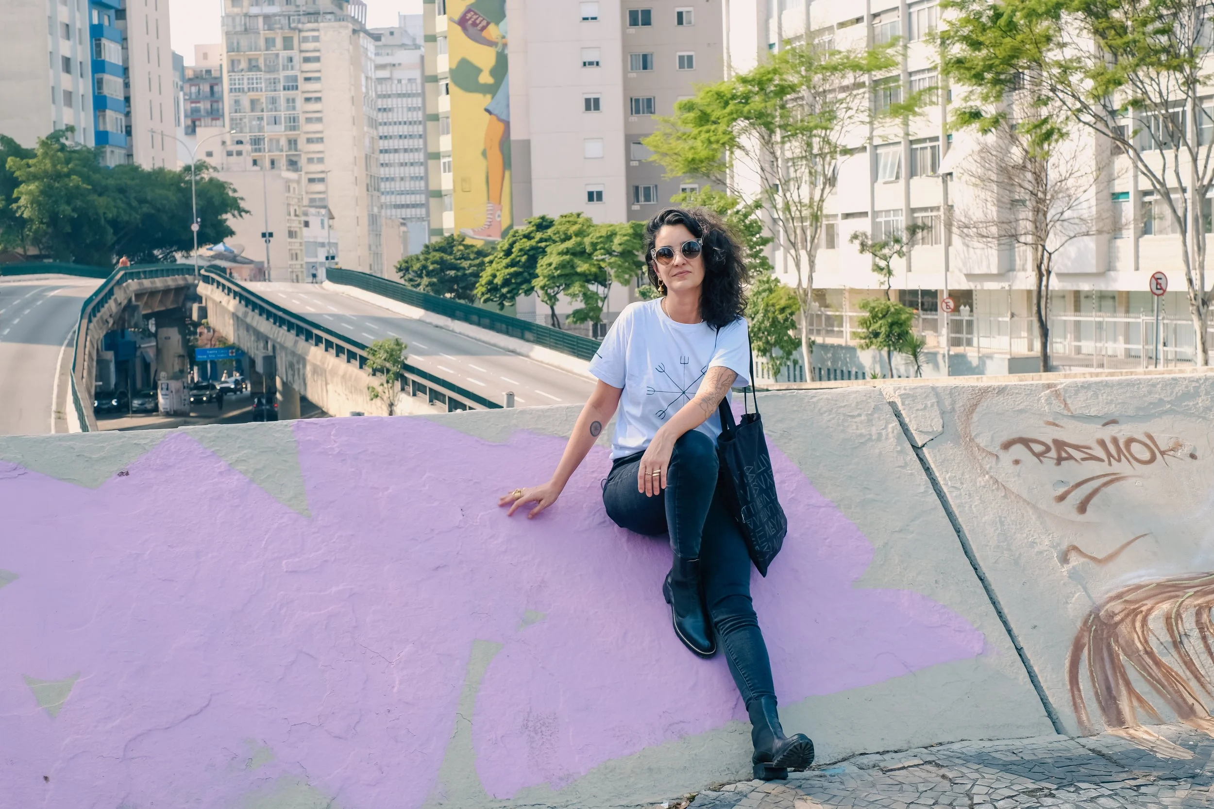 A woman with black curly hair, sunglasses, and tattoos sitting on a purple graffitied concrete barrier in an urban area with tall buildings, trees, and a road in the background.
