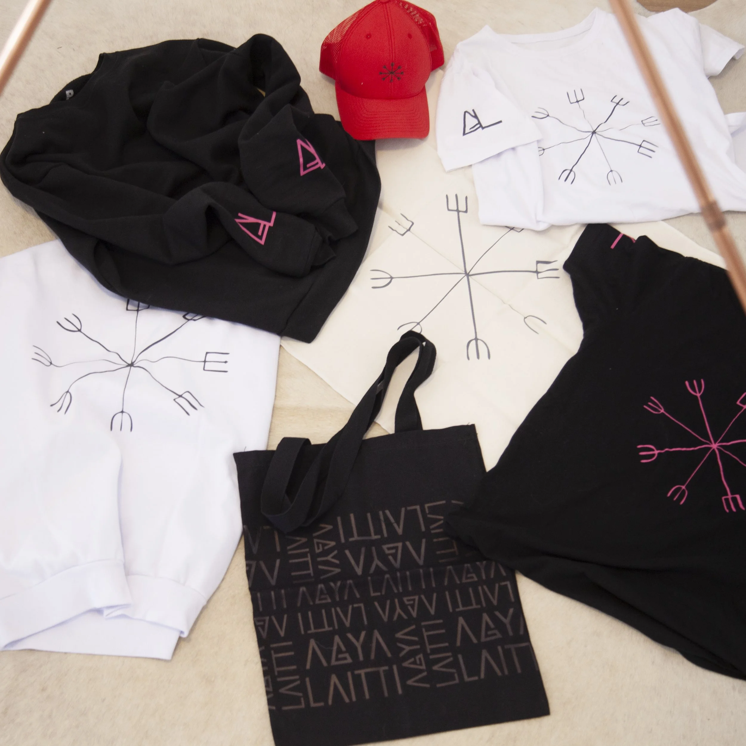 Clothing items featuring Valknut symbols, including black and white shirts and a black hoodie with Valknut designs, a black tote bag with bold Valknut text, and a red cap with a small Valknut symbol set on a beige surface.