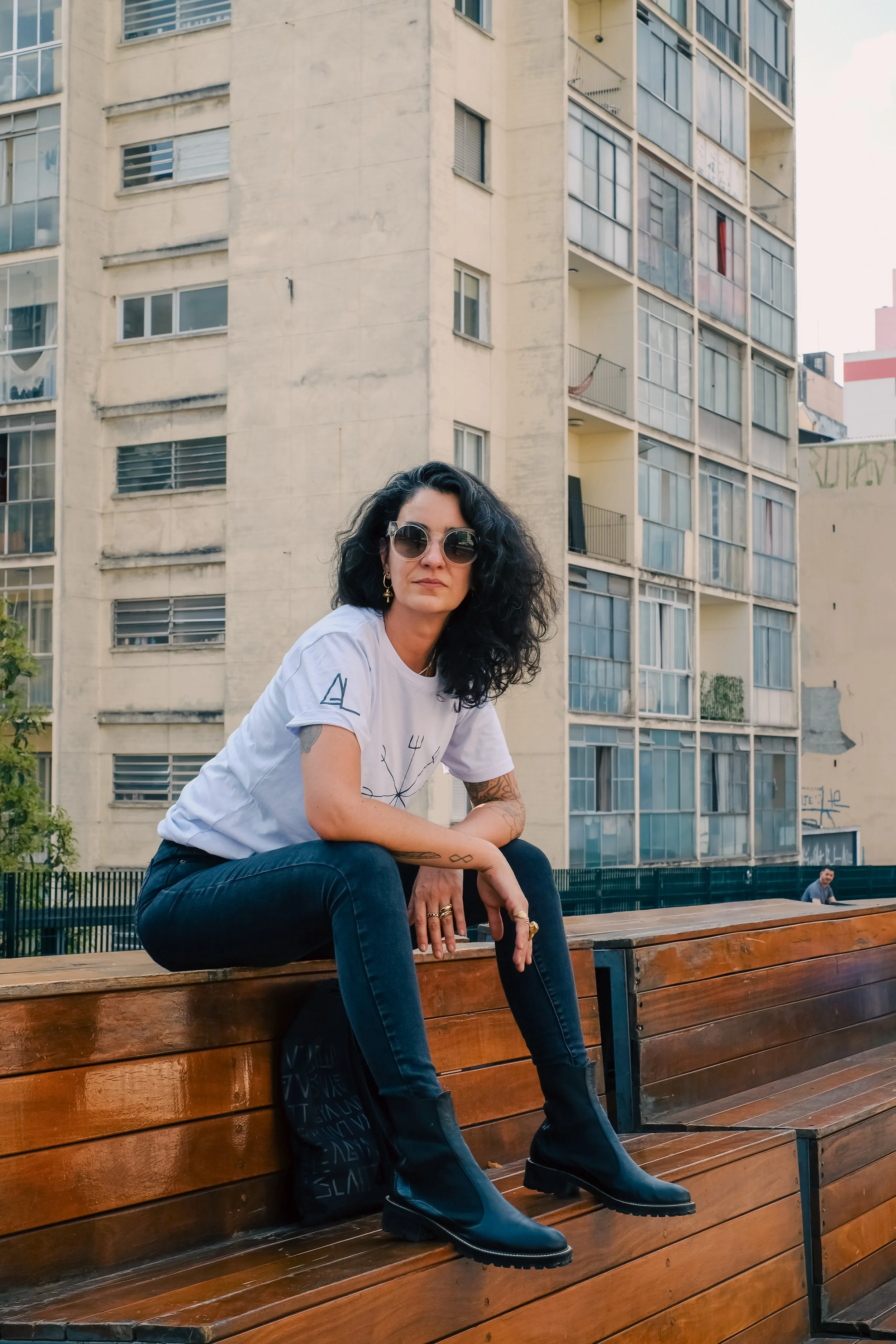 A woman with curly black hair and sunglasses sitting on a wooden bench in an urban setting with tall apartment buildings in the background.