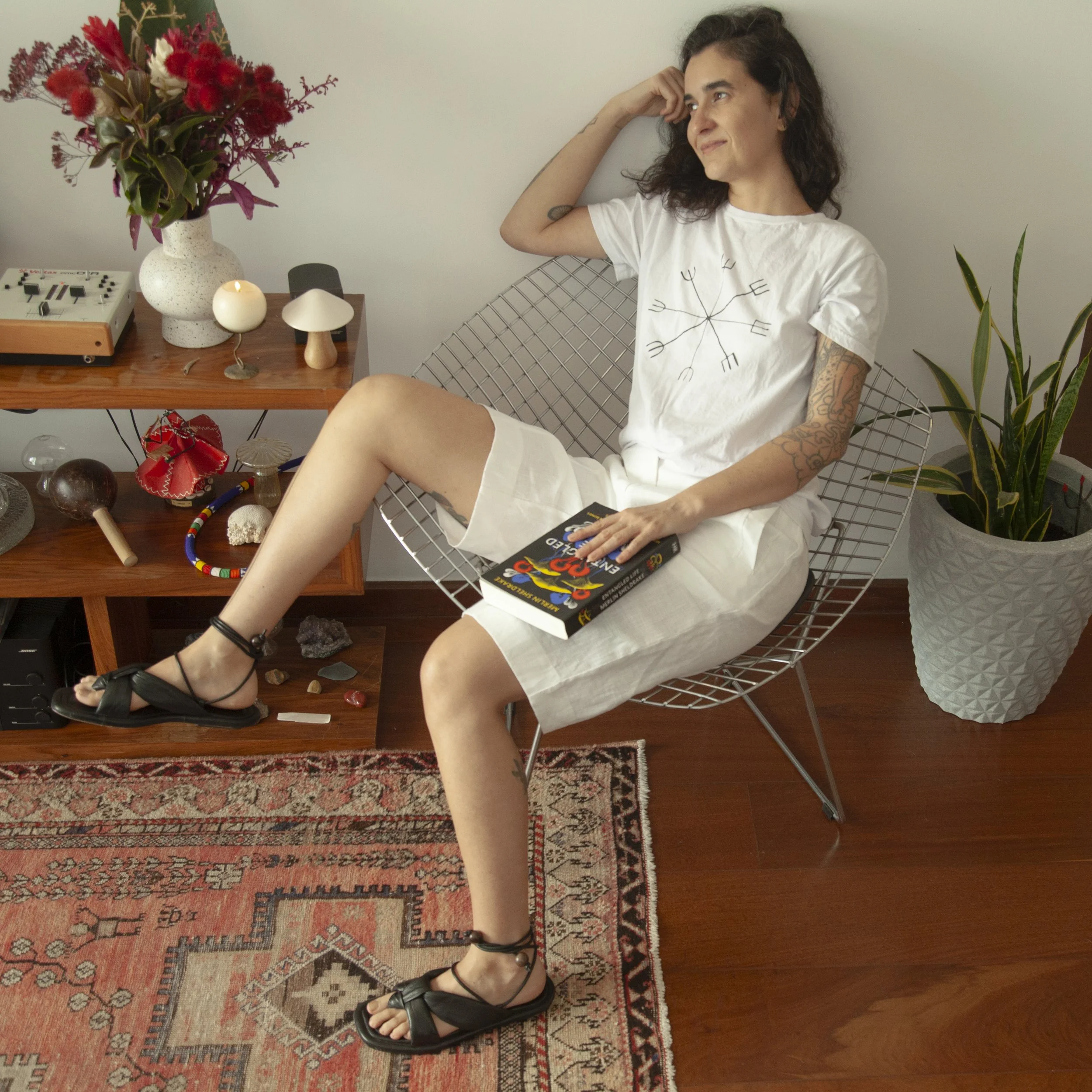 A woman with tattoos sitting sideways in a wire chair, holding a book, wearing a white t-shirt and shorts, with a plant and decorative objects on a wooden shelf nearby.