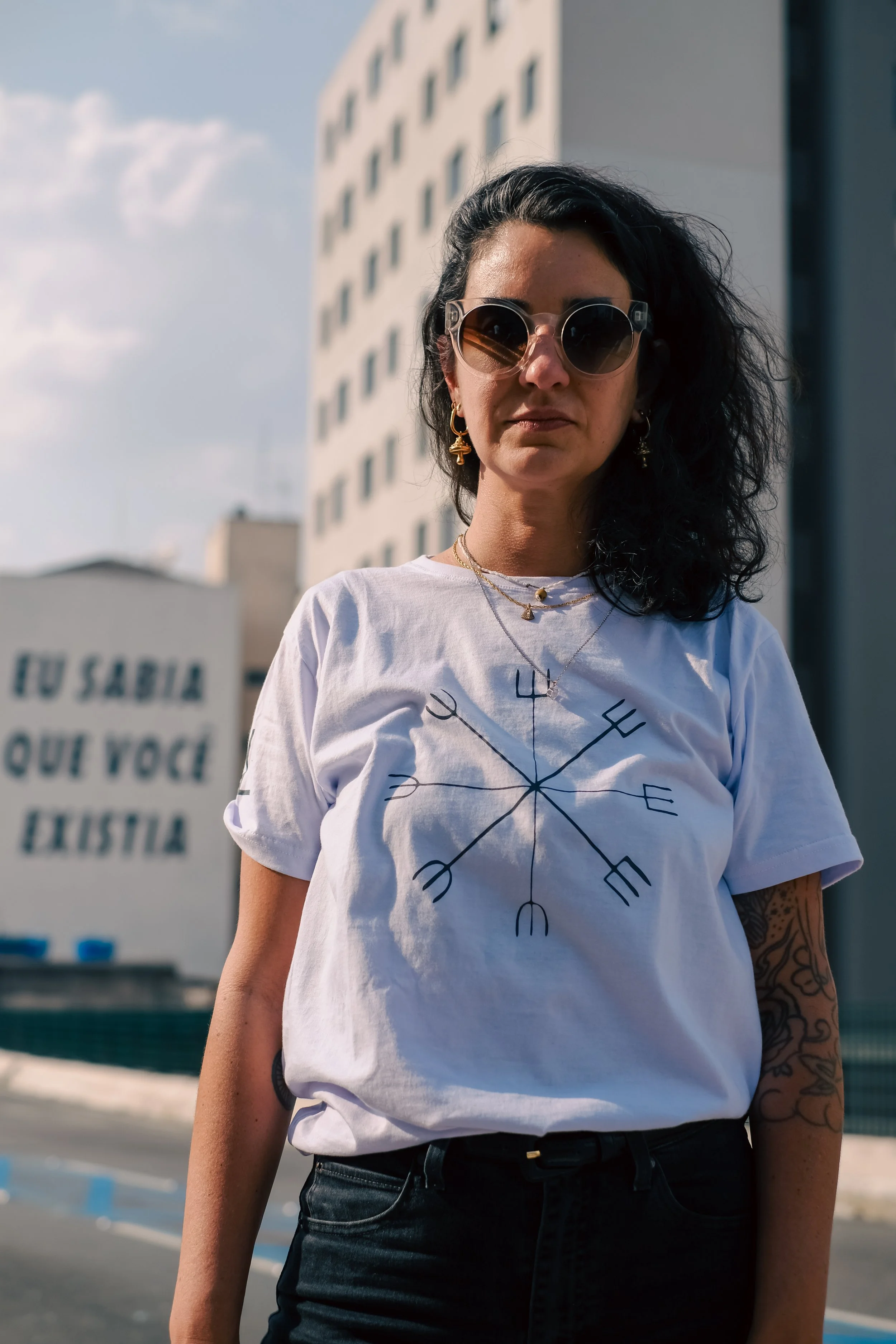A woman with dark, curly hair wearing sunglasses and gold jewelry, standing outdoors in front of a tall building. She is wearing a white T-shirt with a compass design and text, and dark pants.