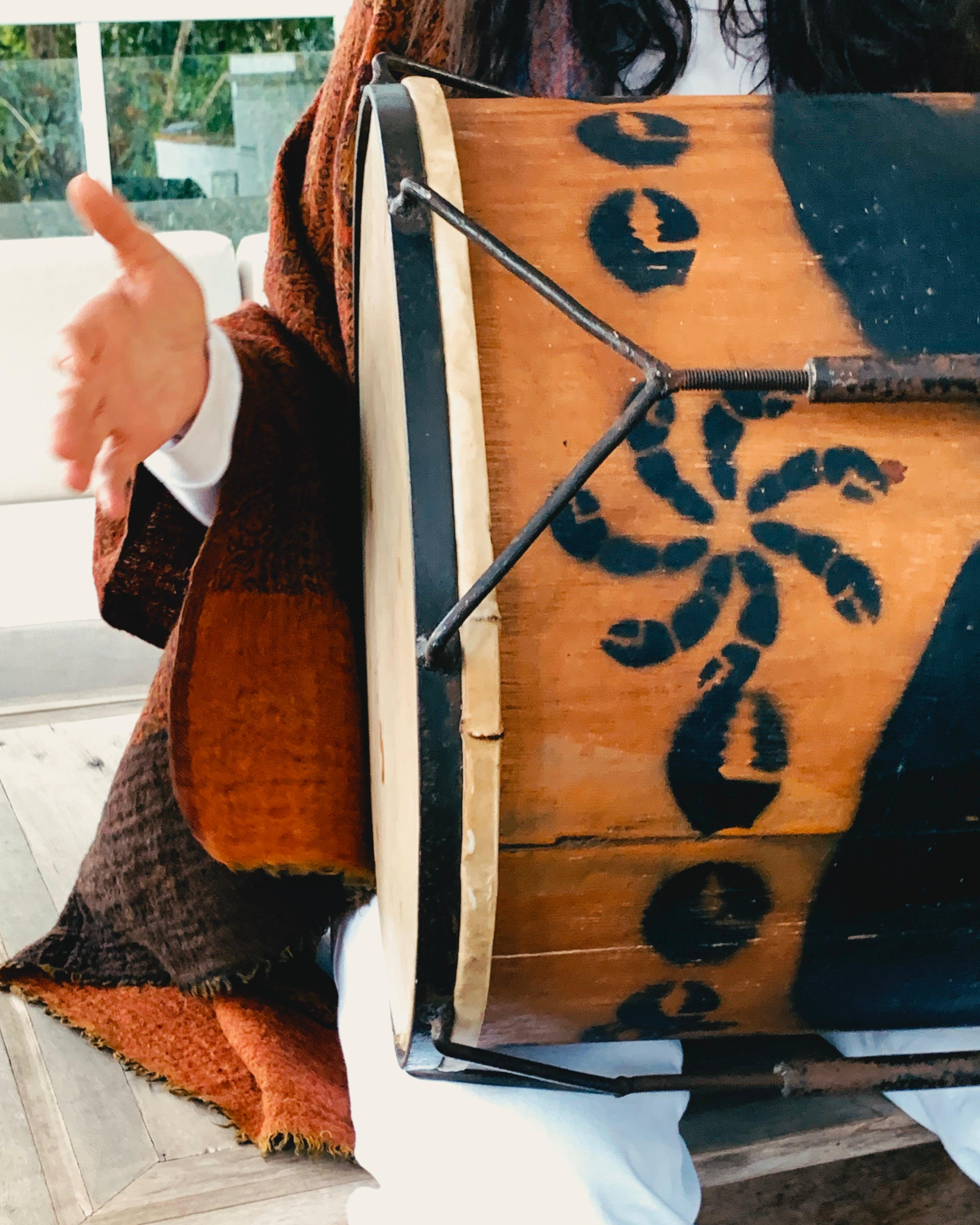A person sitting at a wooden table using a lever device to lift a large pot or container. The table and pot have black decorative patterns. The person's hand is visible, gesturing or holding the lever, with a white sleeve underneath a brownish-red sweater. The background shows a window with outdoor scenery.