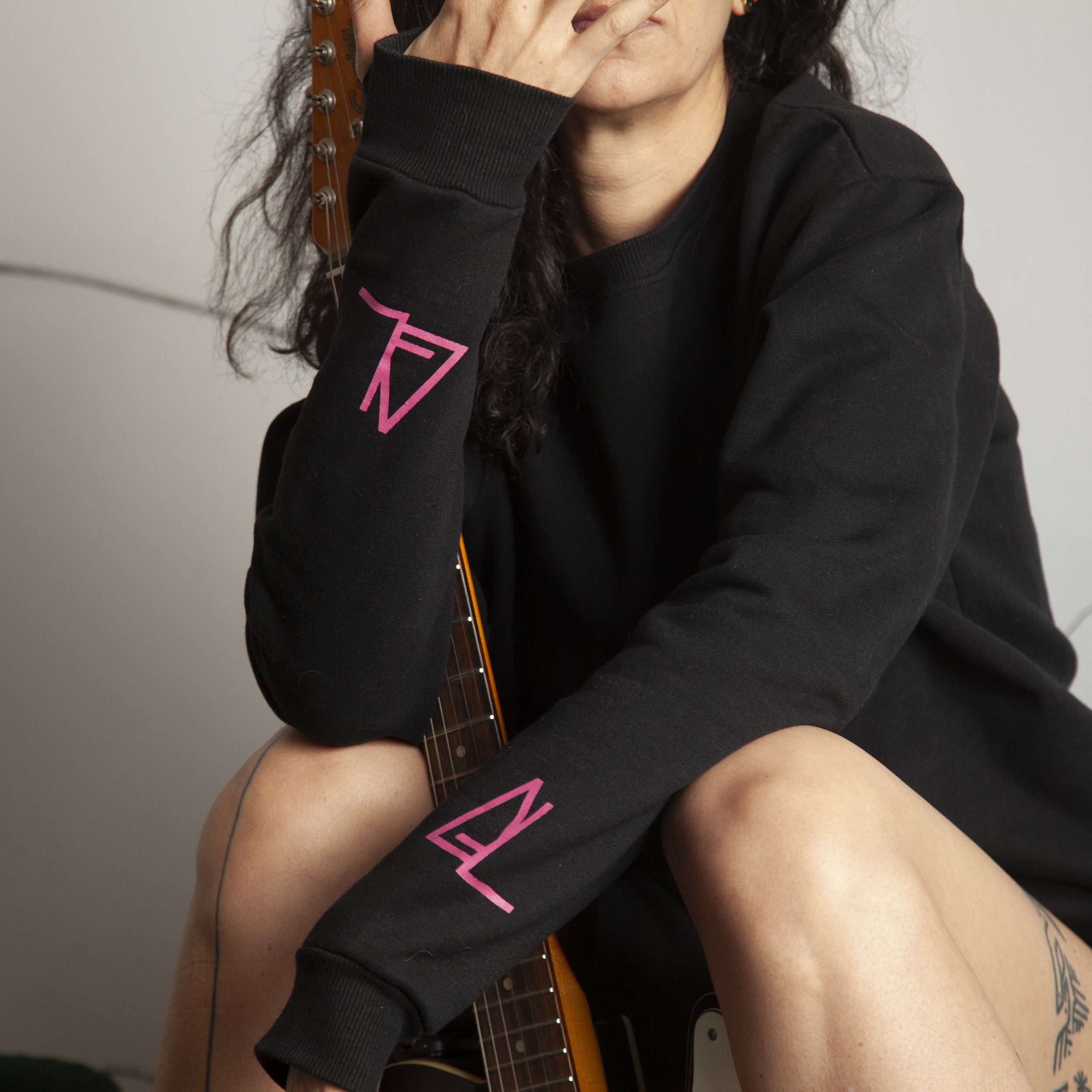 A person wearing a black sweatshirt with pink logos on the sleeve, sitting on a chair and holding an electric guitar, with a tattoo visible on their leg.