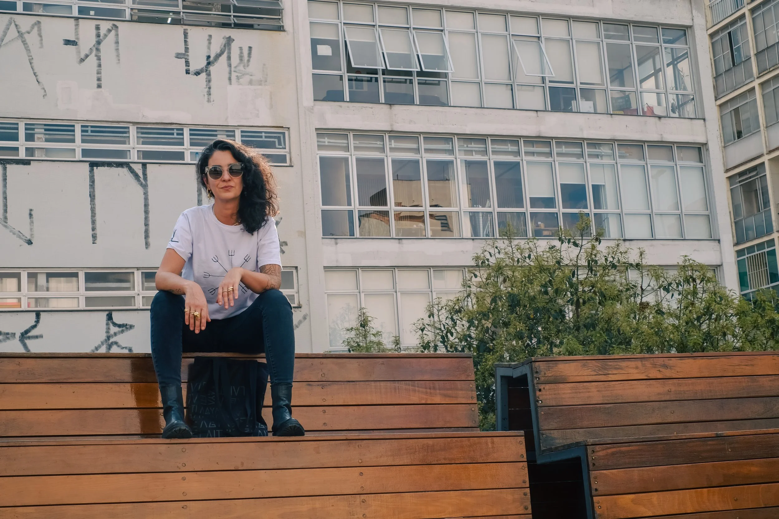 A woman with curly black hair, wearing sunglasses, a white T-shirt, and black pants, sitting on a bench outdoors in an urban area with apartment windows and a building in the background.