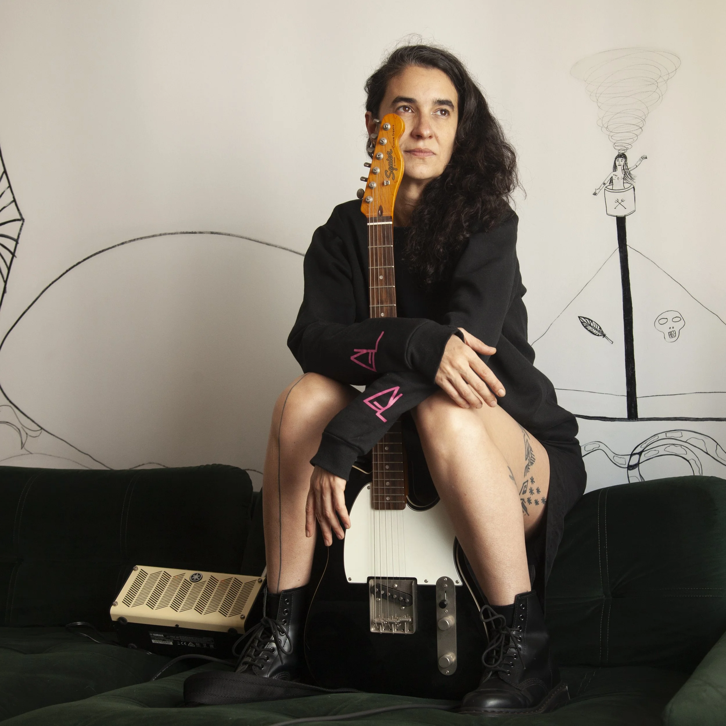 A woman with long curly dark hair sitting on a black couch, holding an electric guitar with the body on the ground and her right arm resting on it, wearing a black jacket and boots. The background features a white wall with hand-drawn black illustrat