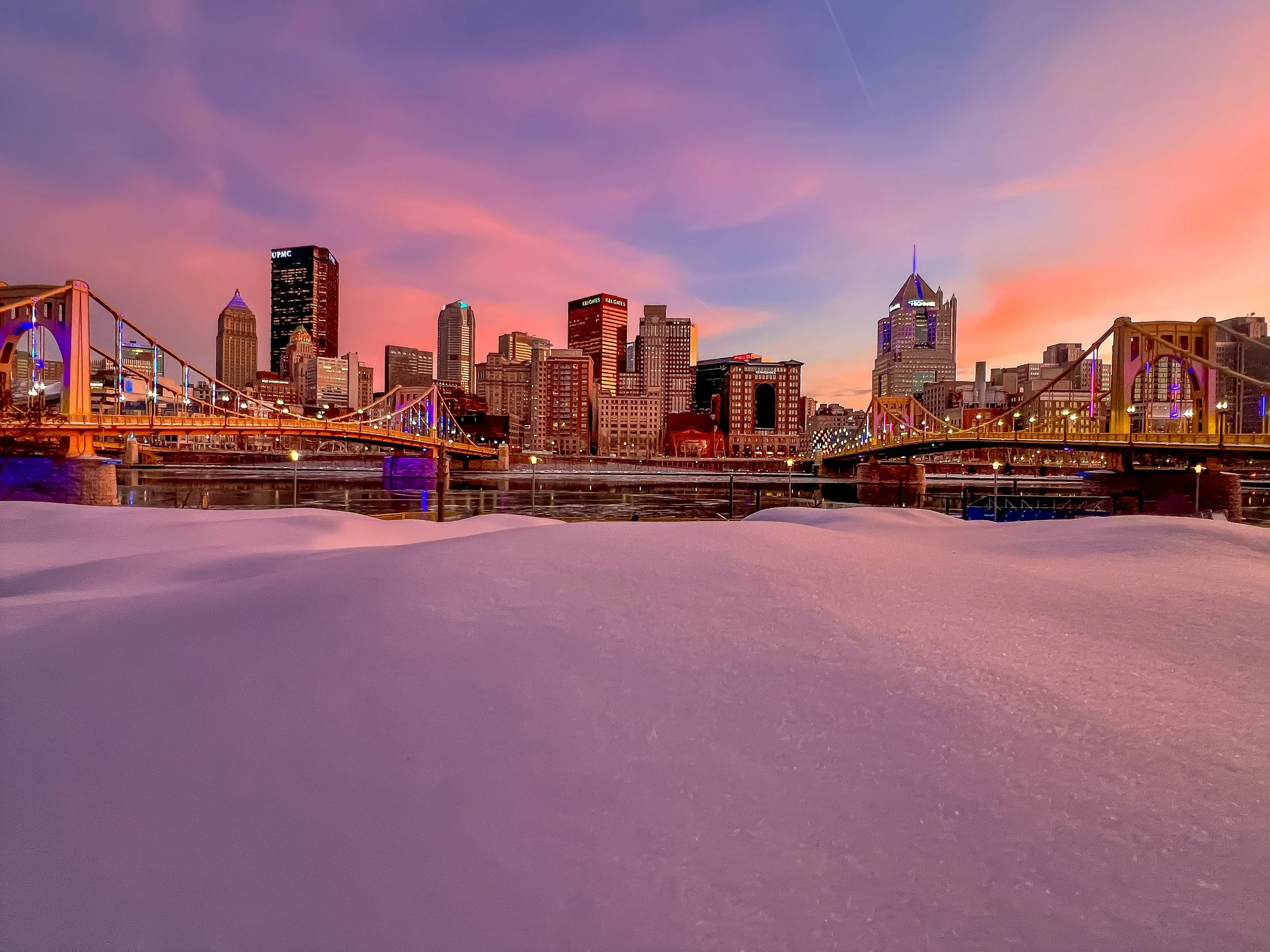 Pittsburgh skyline sunset framed by bridges and river during winter snow with pastel sunset sky.