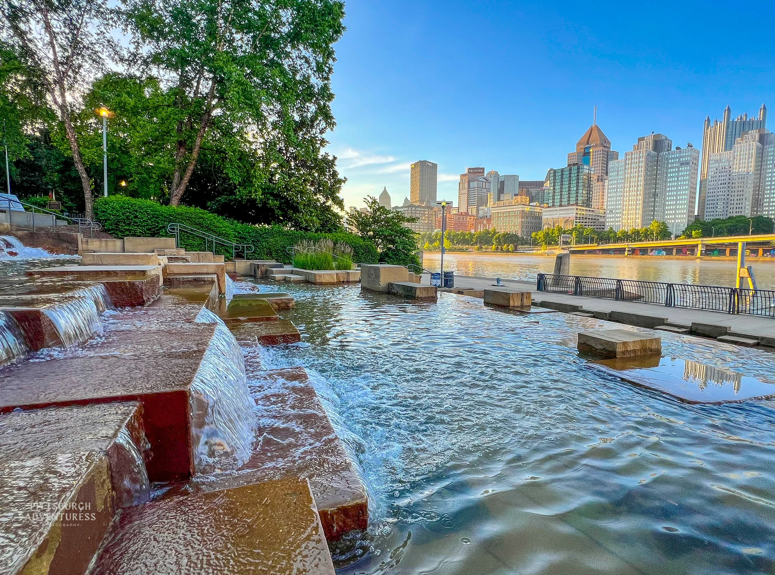 Pittsburgh skyline viewed from riverfront  water steps with flowing water and trees in the foreground.
