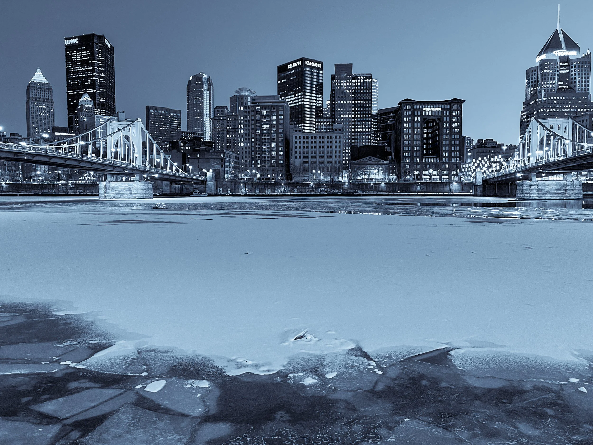 Pittsburgh skyline in winter with snow-covered river and monochrome cityscape.