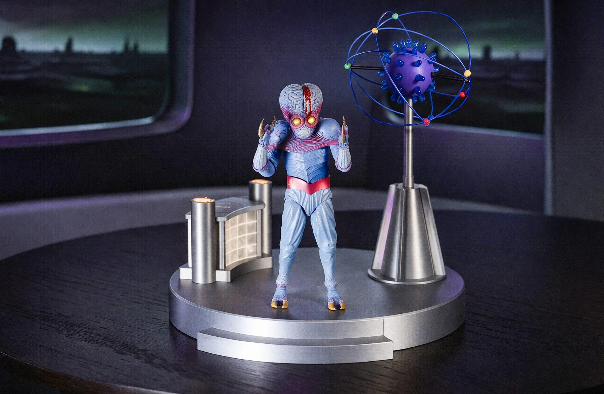 A detailed figurine of a humanoid alien with glowing pink eyes, blue skin, and a large brain head, standing on a circular platform with a sci-fi style background. The figure has hands near its head, horns on its shoulders, and is wearing a red belt and light blue suit, with a large orbital device next to it.