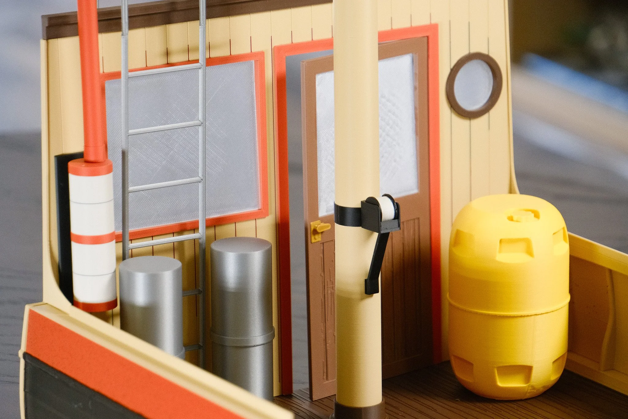 Close-up of a miniature yellow building with a brown door, a yellow propane tank, silver drums, and orange and gray safety equipment on the wall.