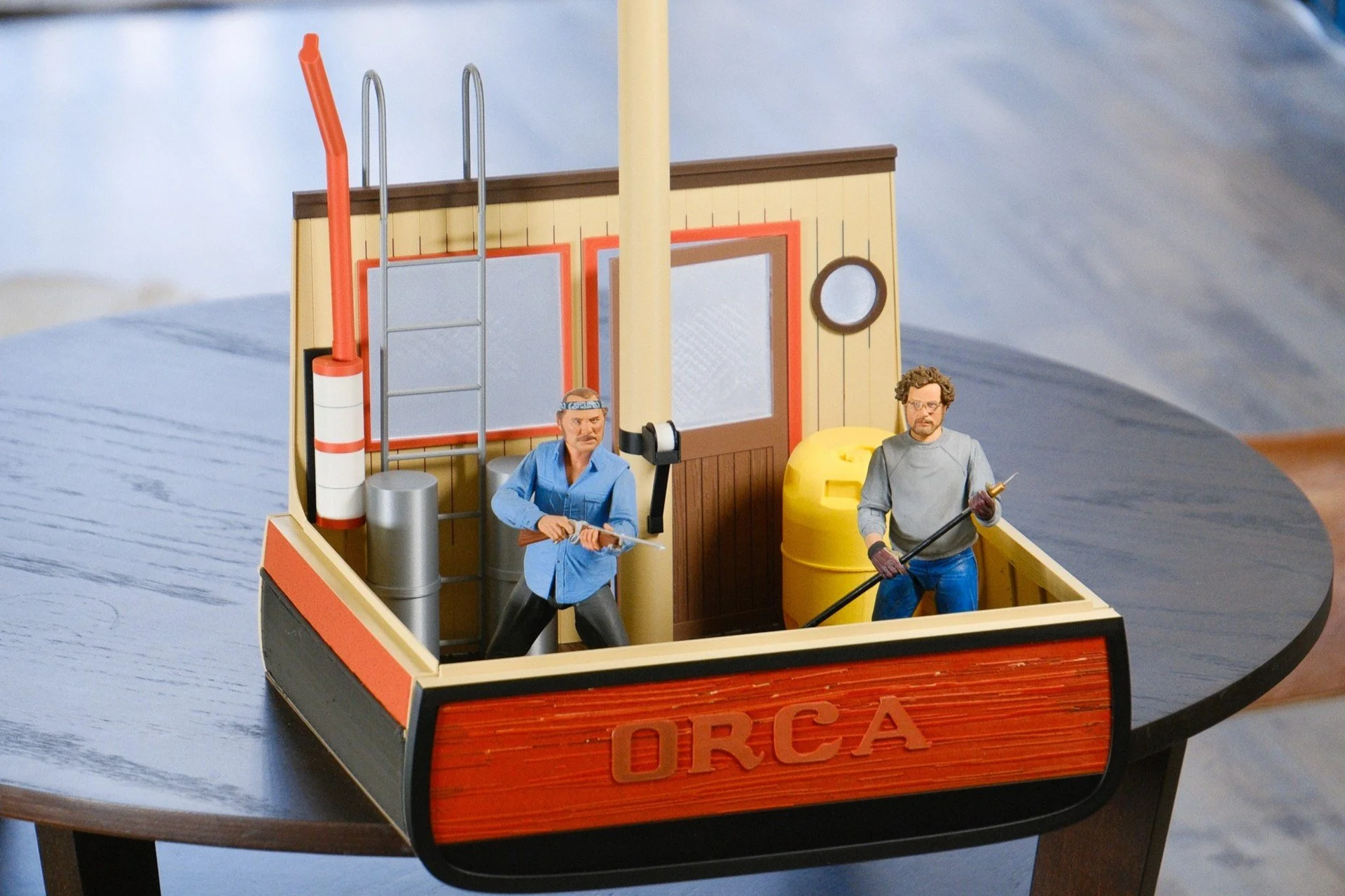 Miniature model of an orca research boat with two figurines inside, one holding a spear and the other with a paddle, on a wooden surface.