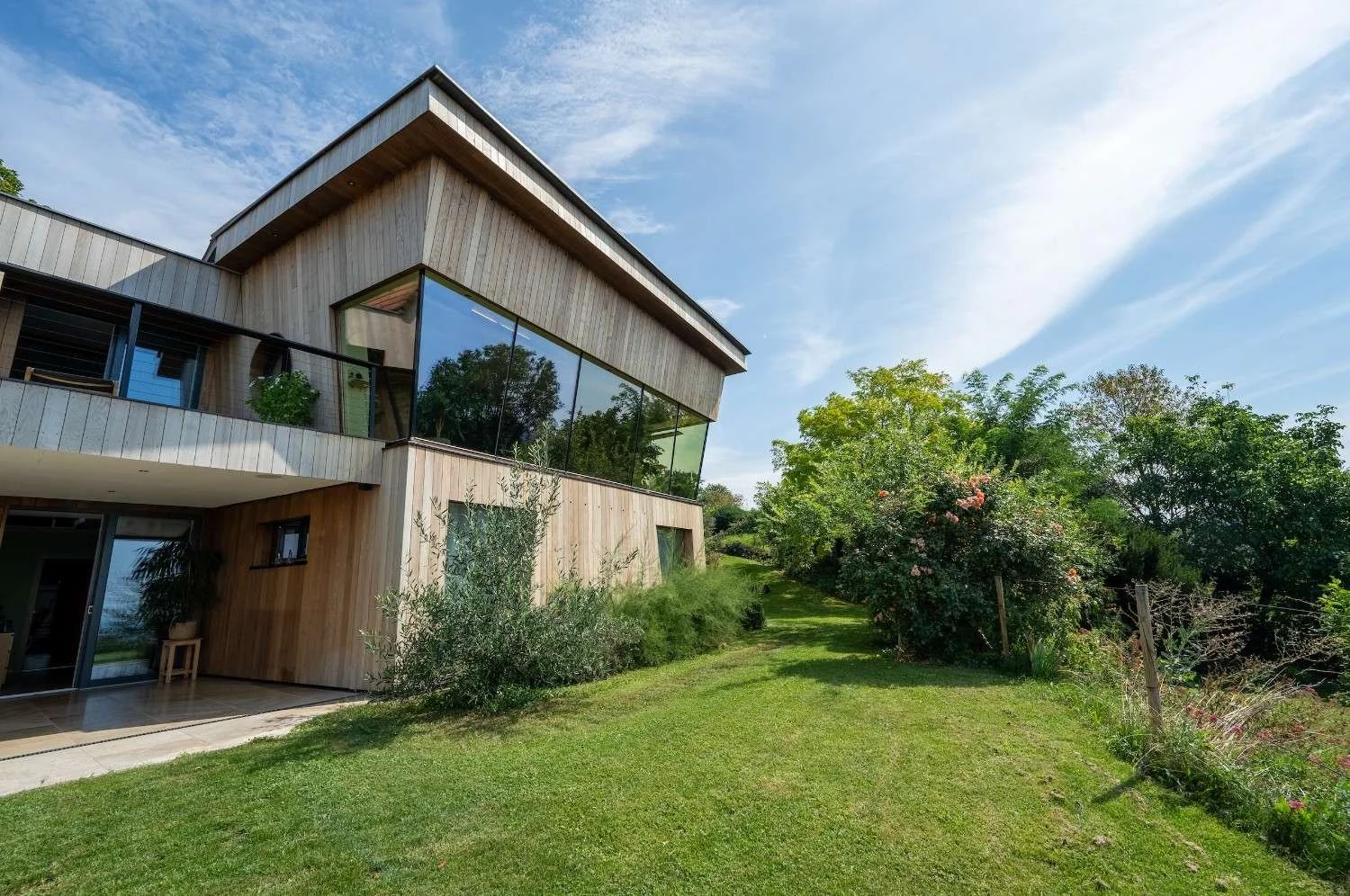 Moderne Holzhaus mit großem Glasfenster vor grünem Garten und blühenden Sträuchern bei blauem Himmel