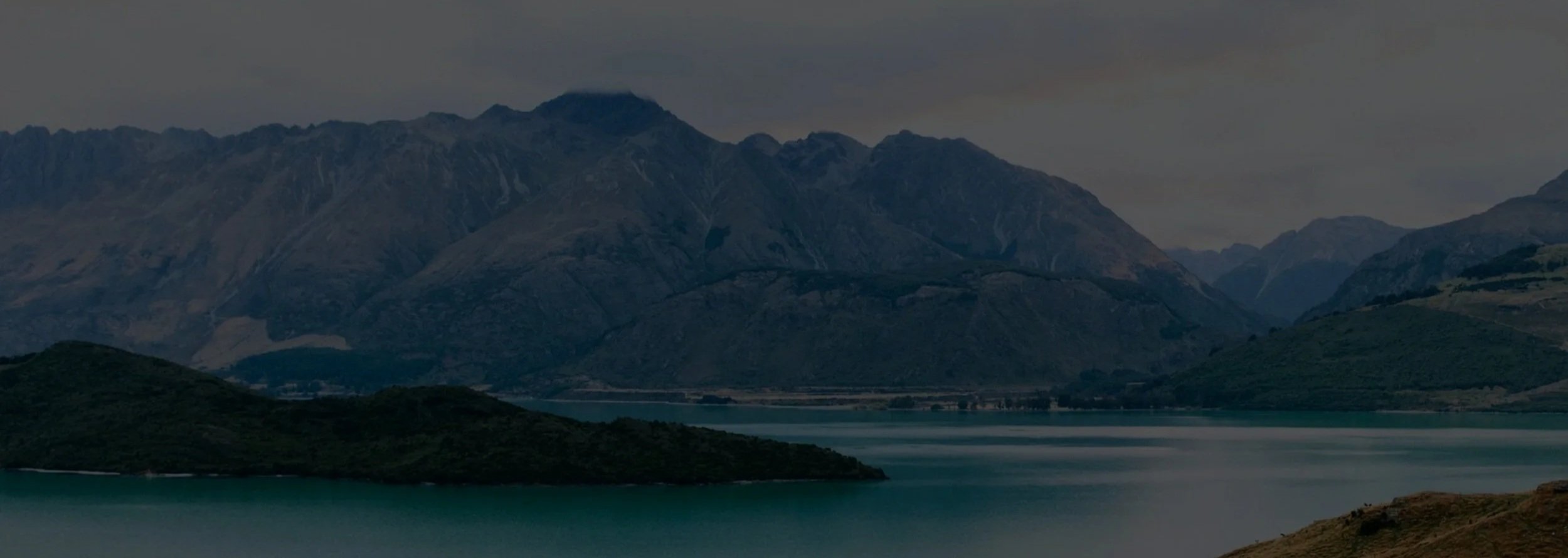 Scenic landscape with a turquoise lake in the foreground, surrounded by green hills, and a range of rugged gray mountains under a cloudy sky in the background.