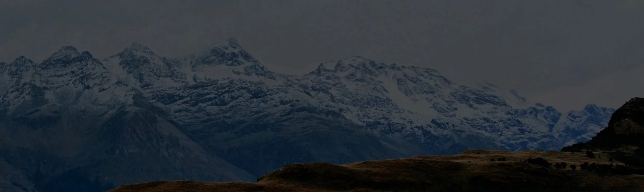 Snow-capped mountain range under cloudy sky with grassy hills in the foreground.