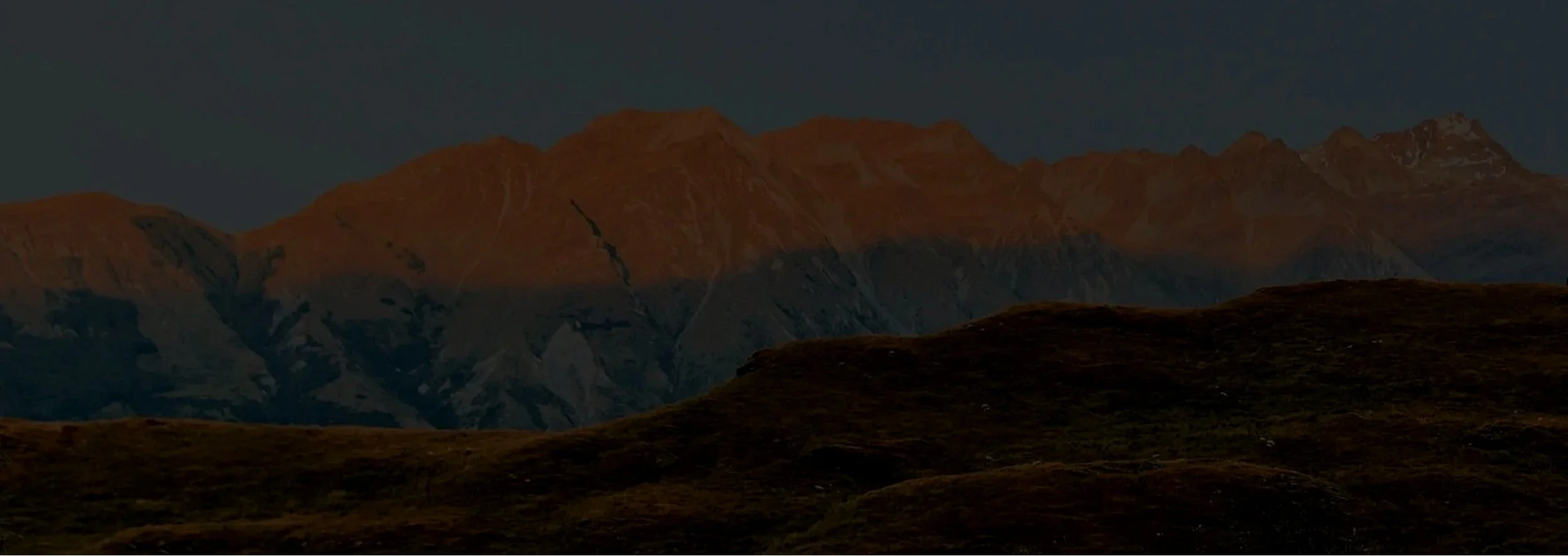 Mountain range with grassy hills in the foreground and a clear sky above, during sunset.