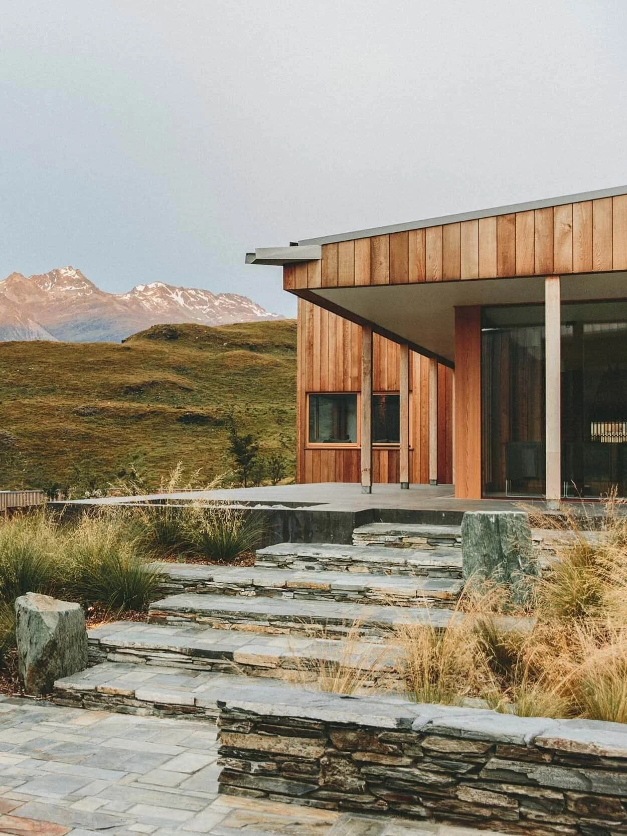A modern house with wooden exterior, large glass windows, and gray stone steps leading up to a patio, set against a landscape of grassy hills and snow-capped mountains in the distance.
