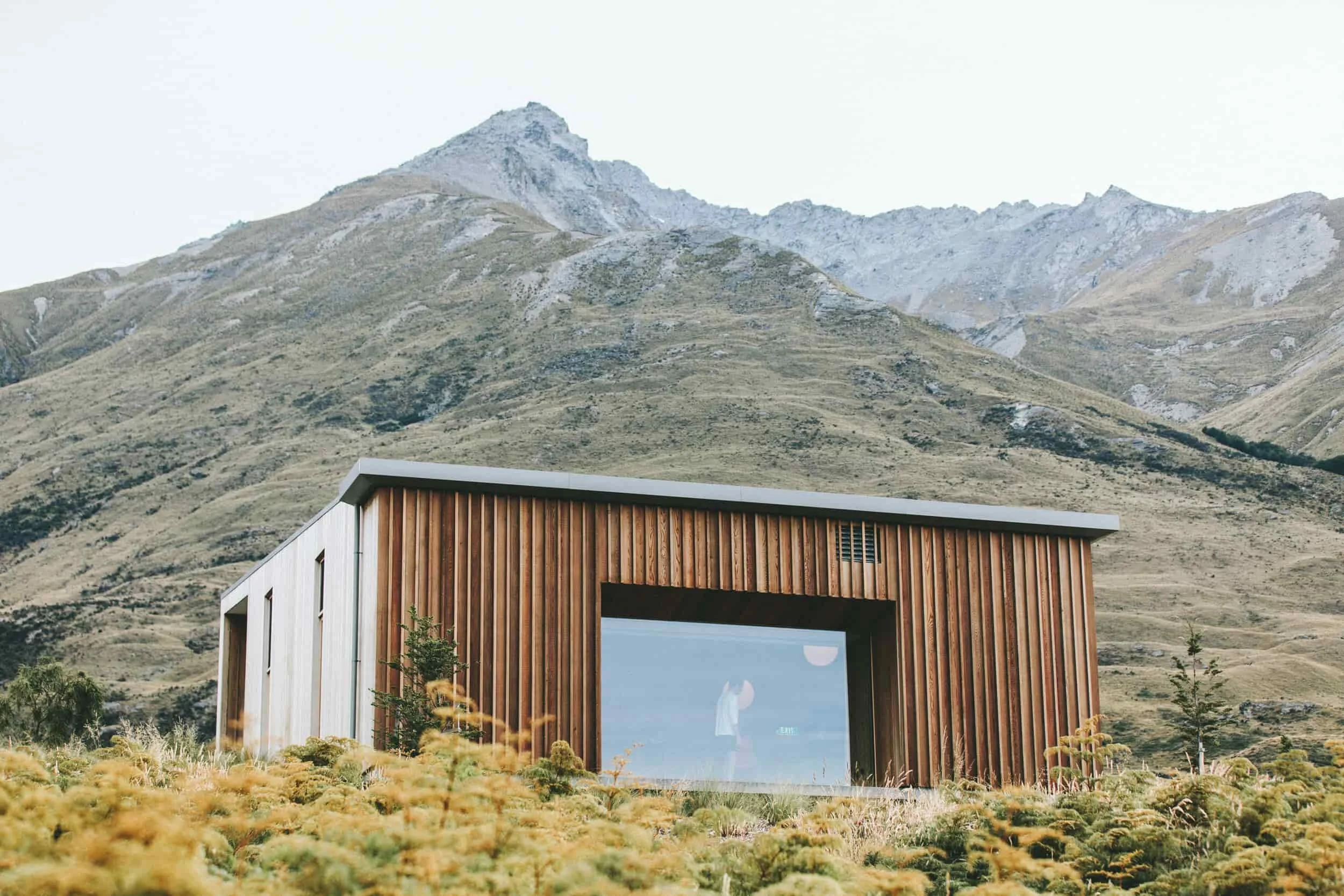 Modern wooden house with large glass window in mountainous landscape with dry grass and sparse trees.