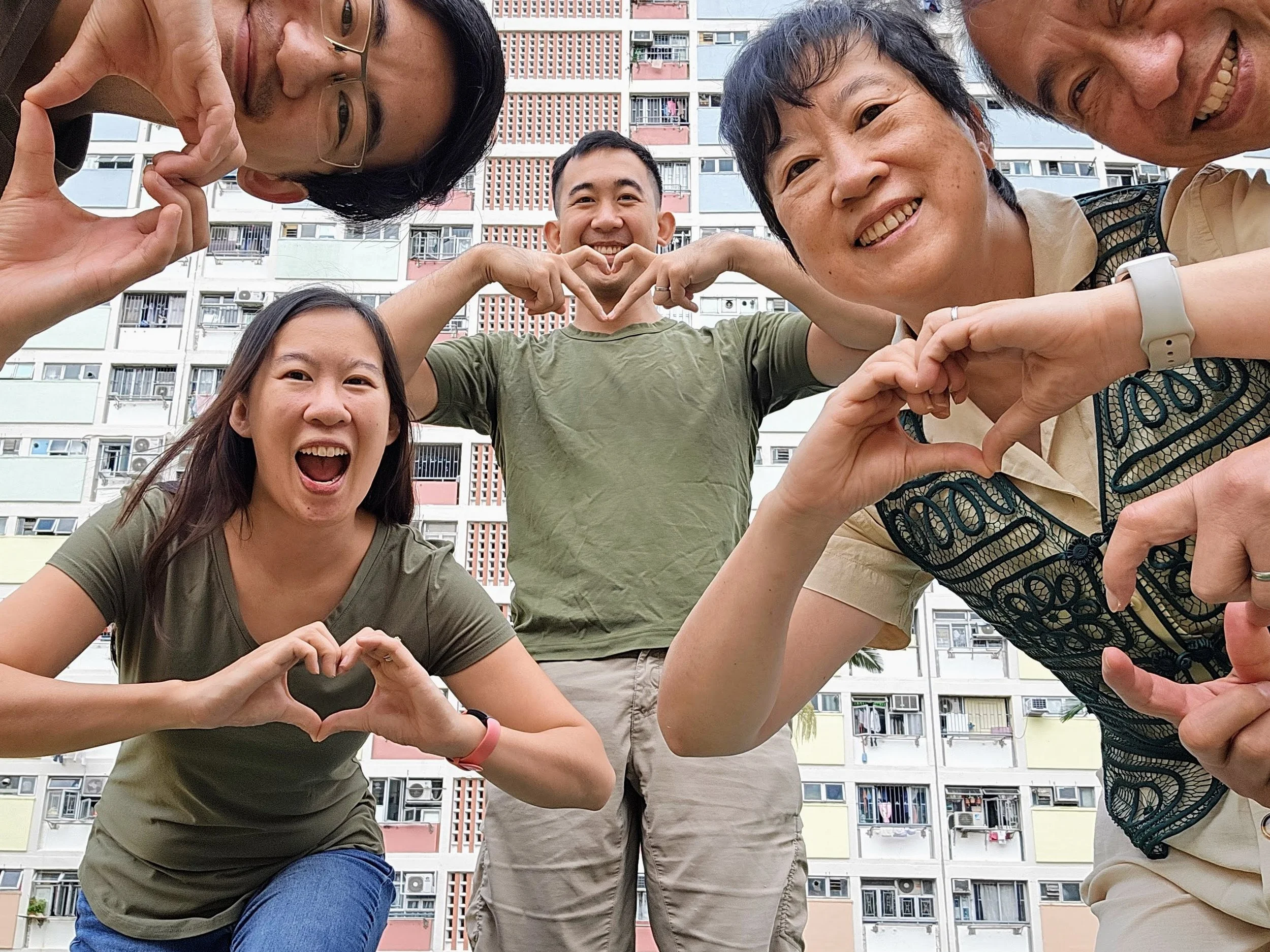 Group of five friends outdoors making various heart shapes with their hands, smiling, with a colorful apartment building in the background.