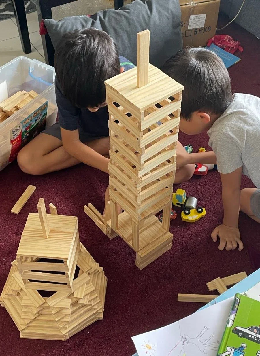 Two children playing with large wooden blocks, building a tall tower and other structures on a red carpeted floor.
