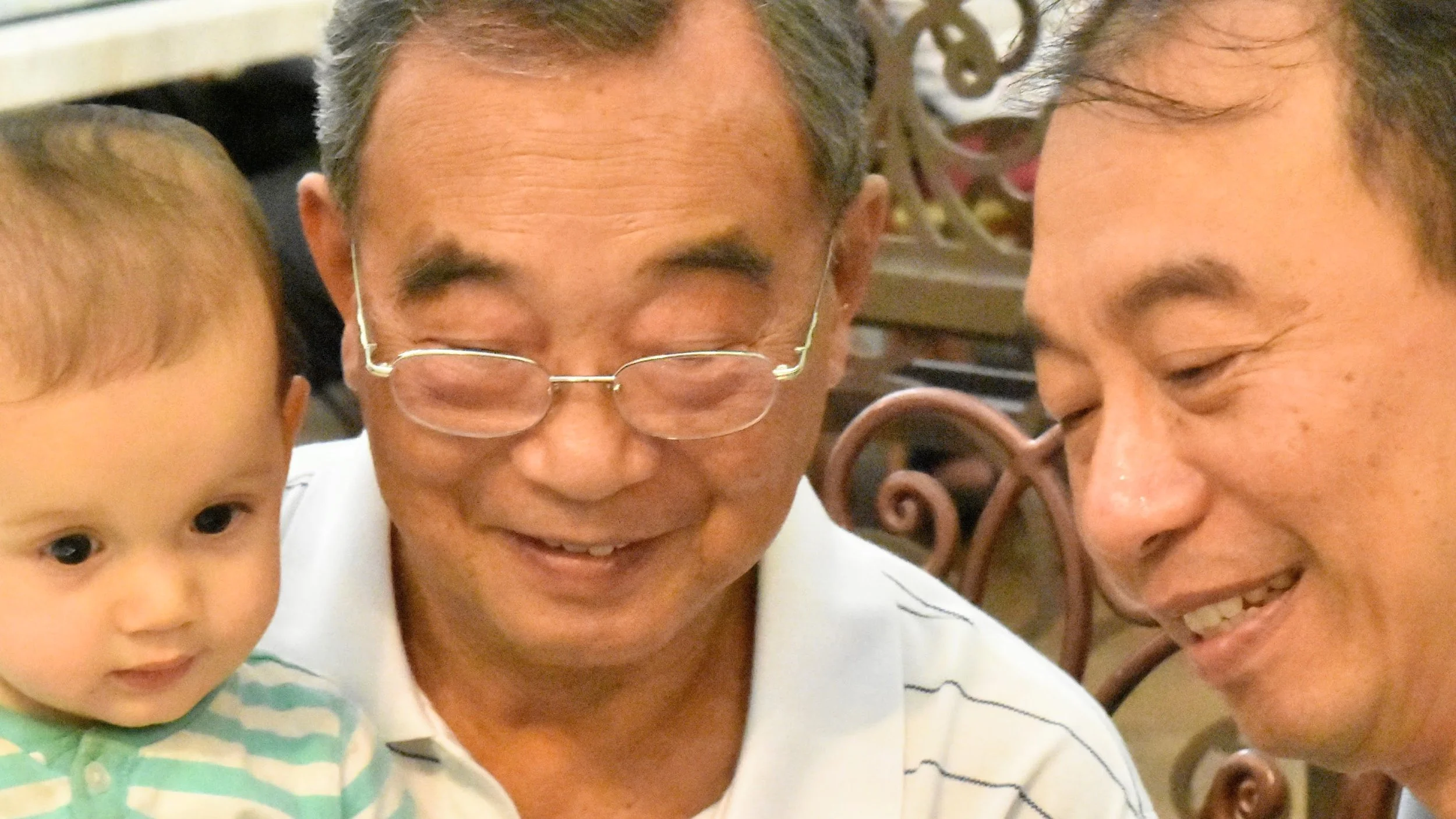 Three people, including a young child and two older men, smiling and looking at a device or object outside the camera's view, in an indoor setting.