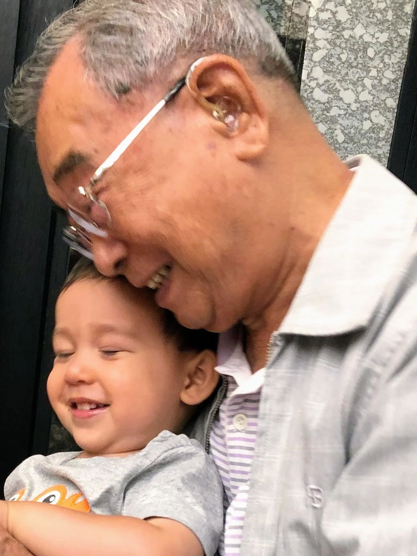 A joyful moment between an older man and a young child, with the man leaning his forehead against the child's head, both smiling with eyes closed.