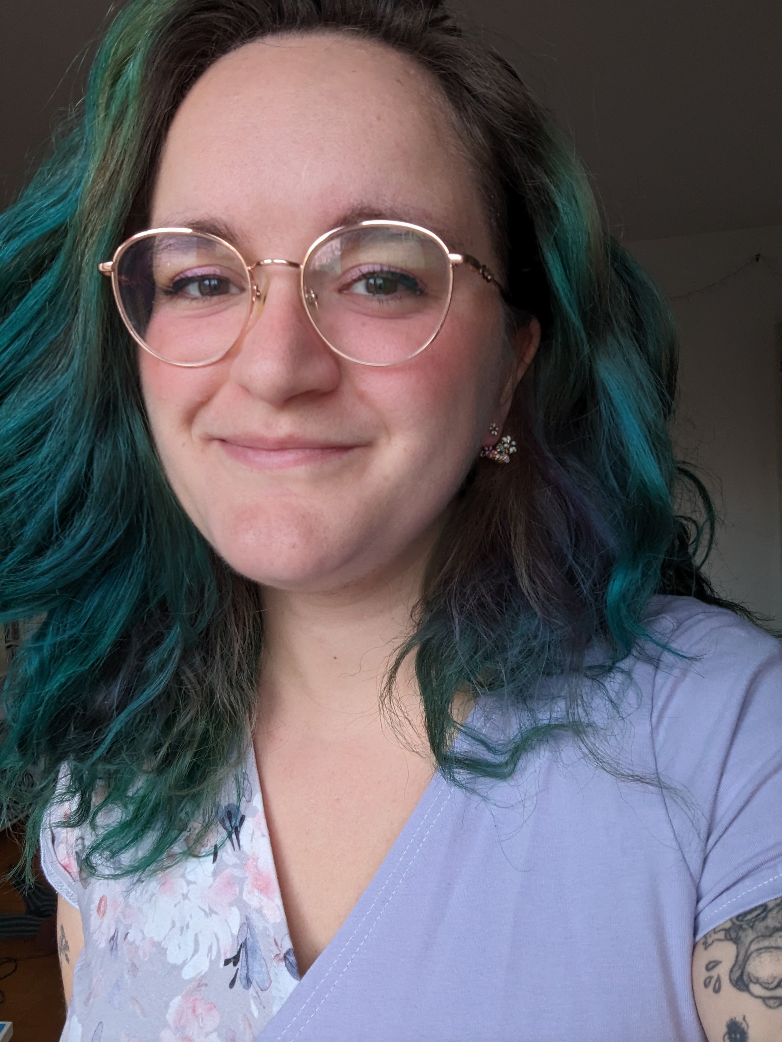 Une femme avec des cheveux longs et bouclés, teintés en vert et bleu, porte des lunettes rondes en métal rose or rose. Elle sourit légèrement, porte des boucles d'oreilles à fleurs et une blouse blanche avec un motif floral, et a un tatouage visible sur son bras droit.