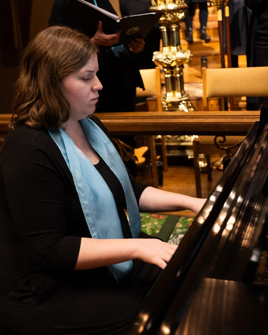 A woman sits at a grand piano with her hands on the keys, eyes closed, wearing black clothes and a long blue scarf