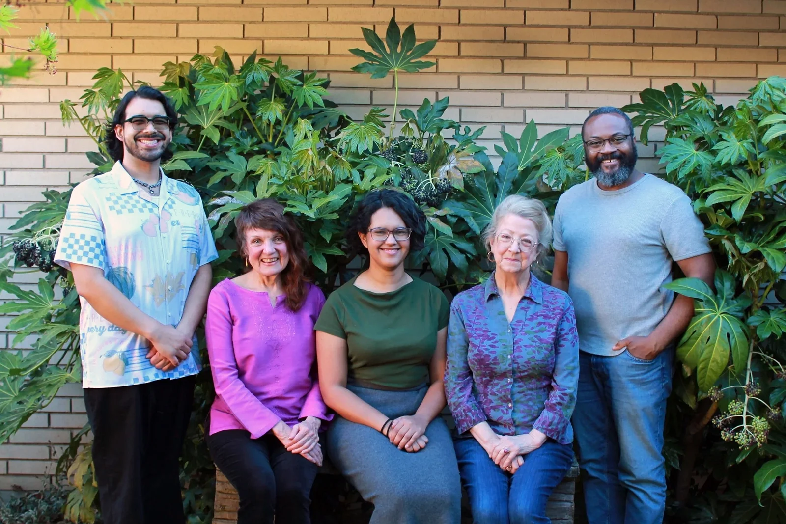 5 people smile at the camera, standing and sitting in front of a plant-covered brick wall