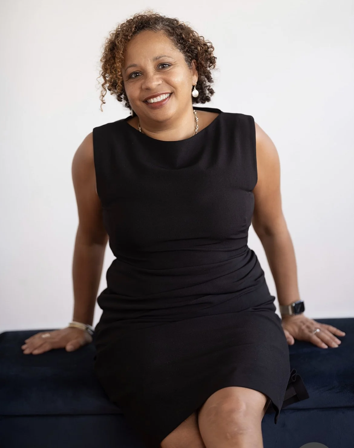 A woman with curly brown hair, wearing a sleeveless black dress, sitting on a dark surface against a plain white background, smiling at the camera.