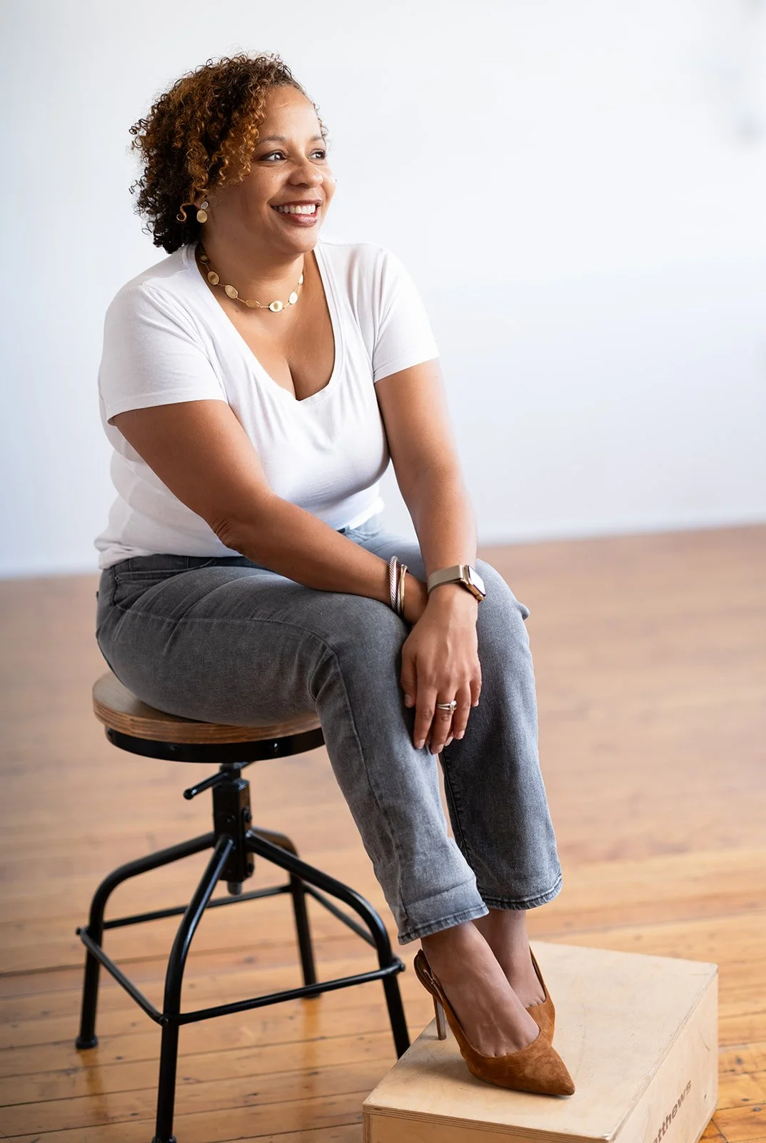A woman with curly hair sitting on a stool, smiling, with her left foot on a wooden box, wearing a white t-shirt, gray jeans, and brown high-heeled shoes, in a room with wooden floor and white wall.