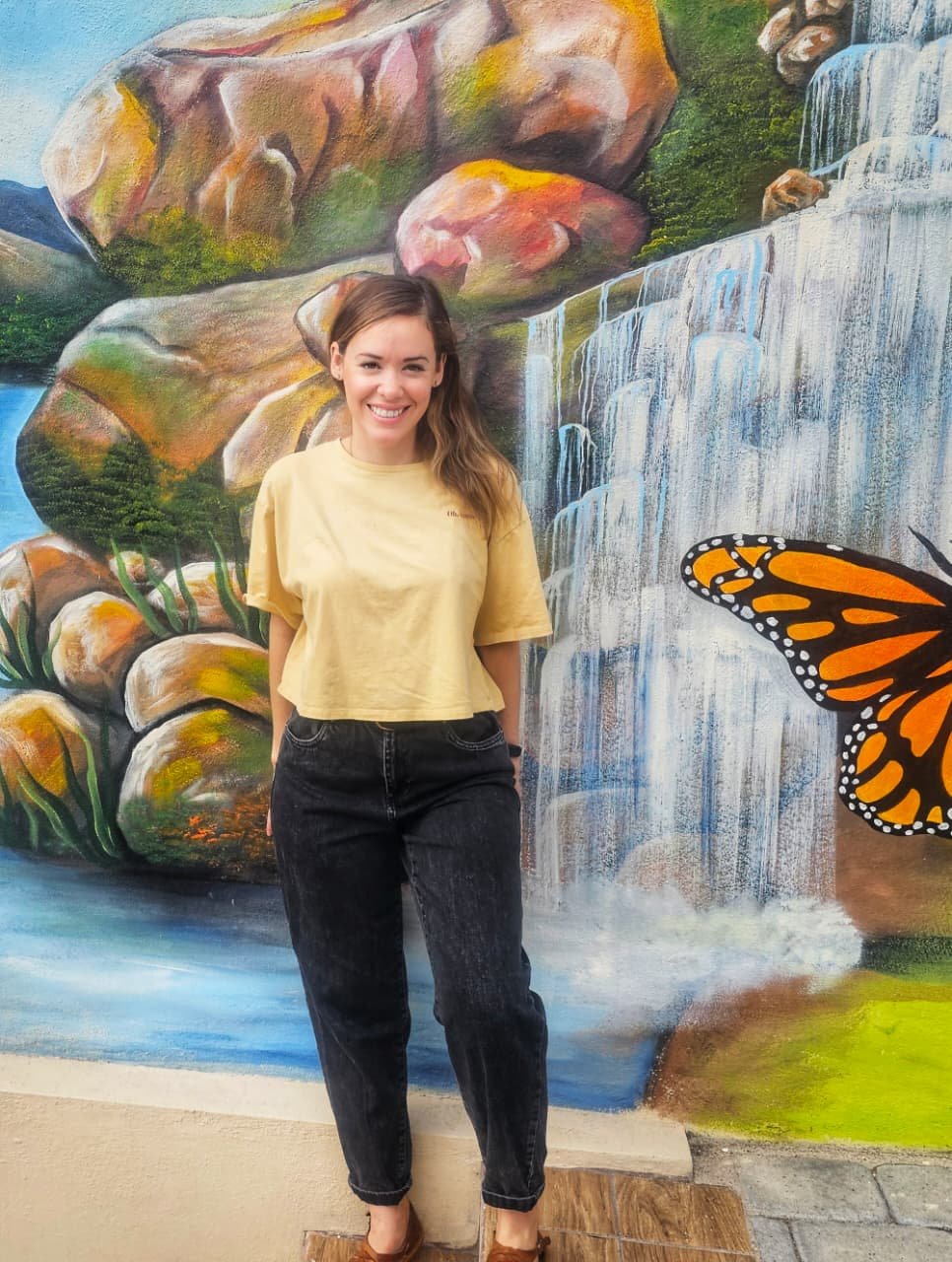 A young woman with long brown hair and a yellow T-shirt standing in front of a colorful mural of rocks, a waterfall, and a butterfly.