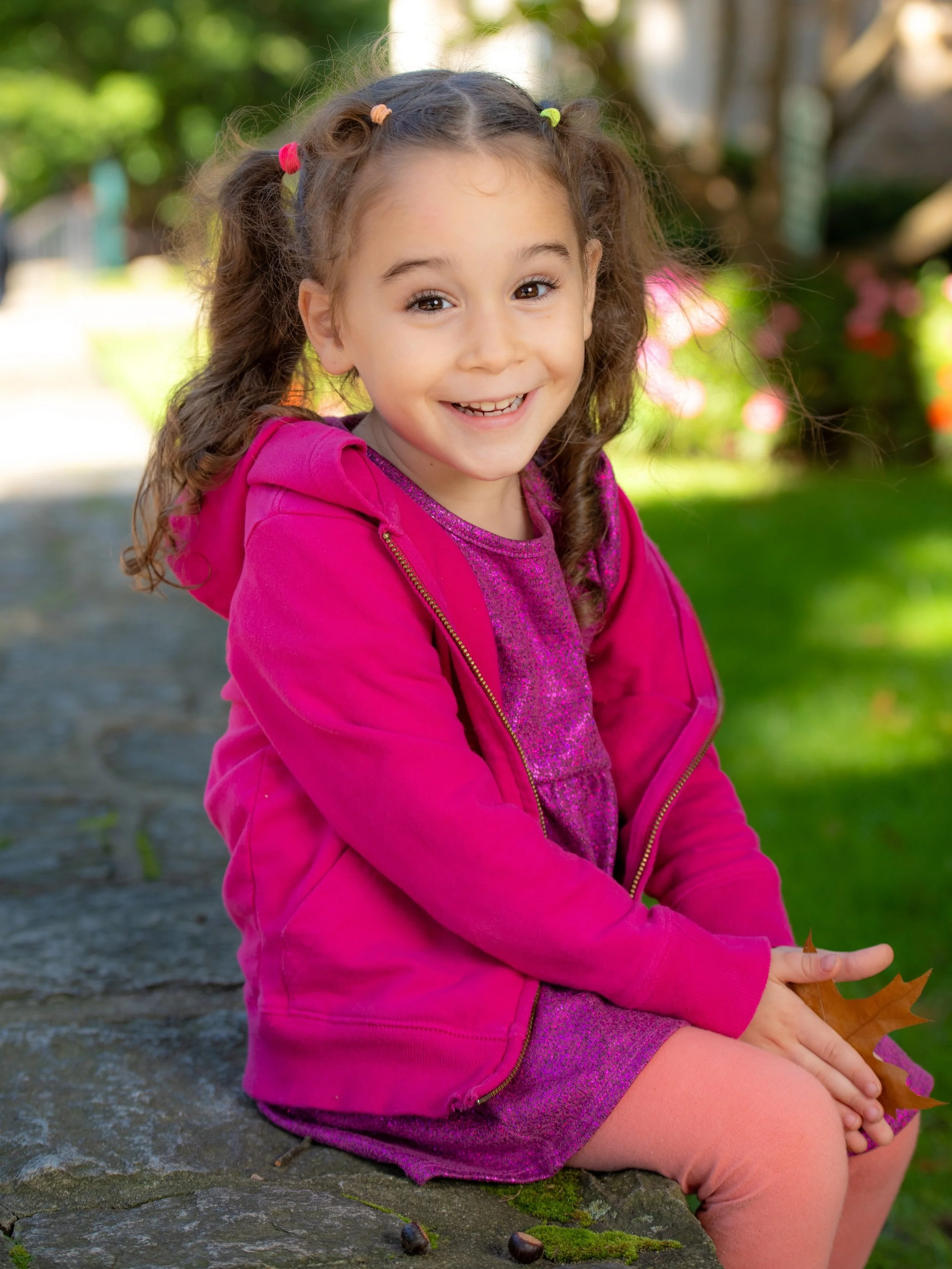 Outdoor school portraits for nursery schools. A smiling young girl with curly brown hair styled in pigtails sitting outdoors on a stone surface in a park with green grass and trees in the background.