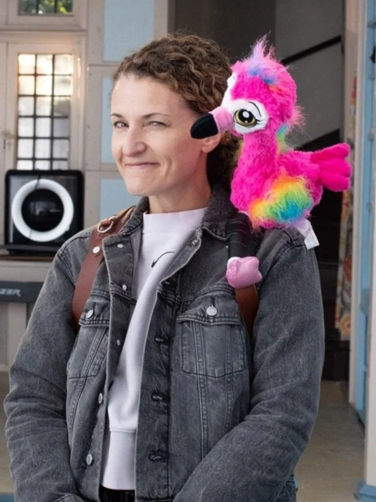 A woman with curly hair wearing a gray denim jacket with a colorful plush pink unicorn toy on her shoulder.