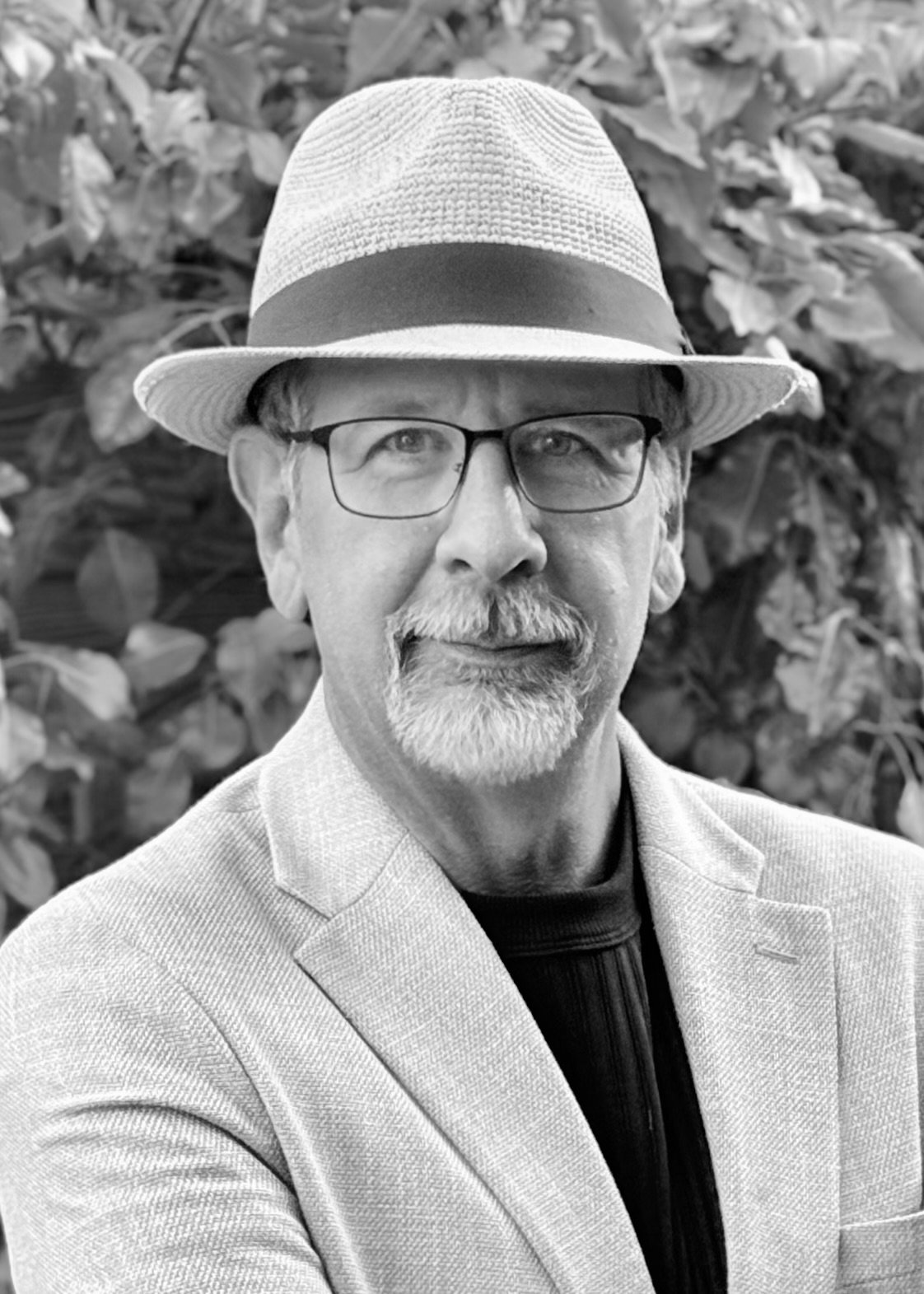 A black and white portrait of a middle-aged man wearing glasses, a fedora hat, a blazer, and a dark shirt, standing outdoors with leafy background.