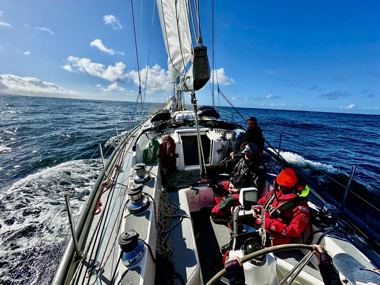 View from a sailing boat with four people onboard under a partly cloudy sky, sailing on the open ocean.