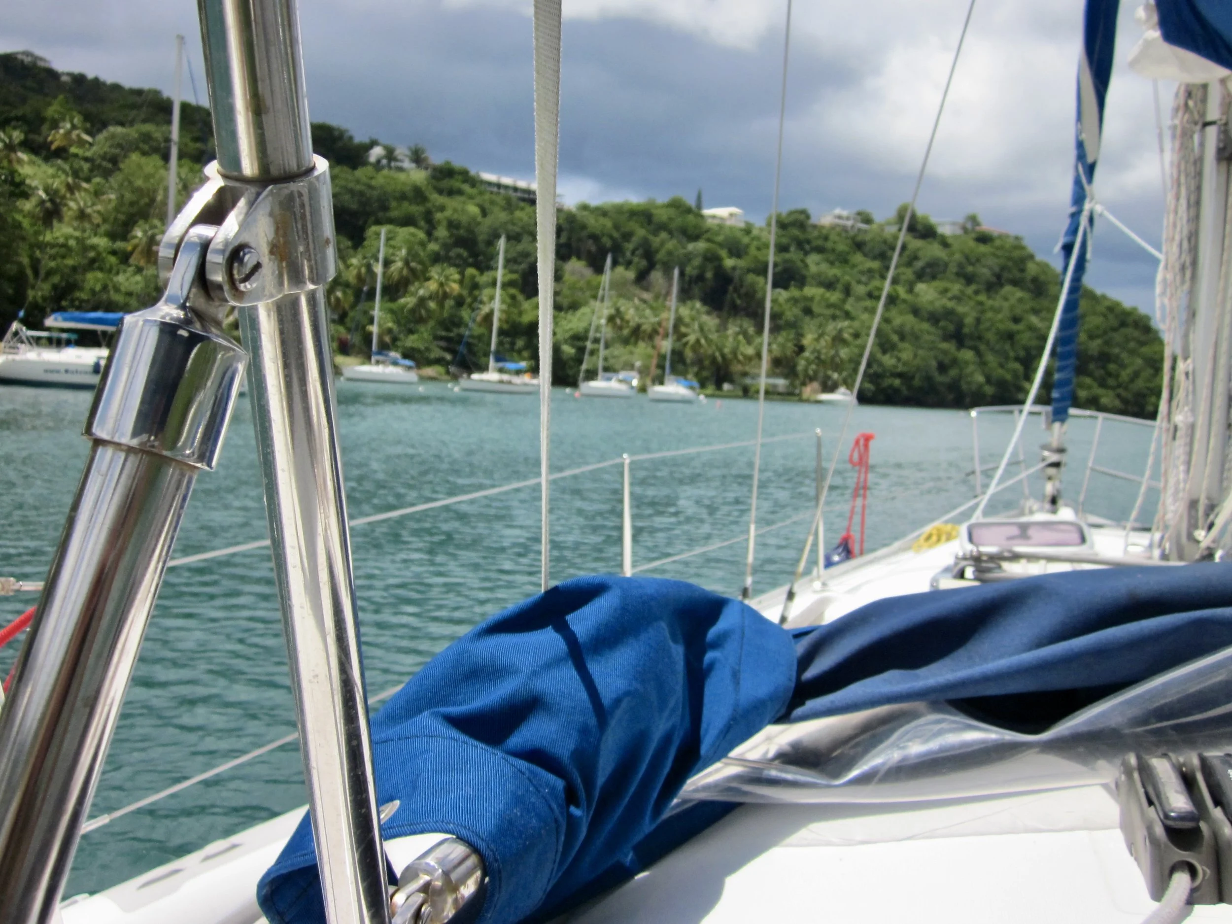 A person wearing a blue jacket sitting on a sailboat, with a focus on metal rigging in the foreground and a lush, green hillside with other boats anchored in the water in the background.