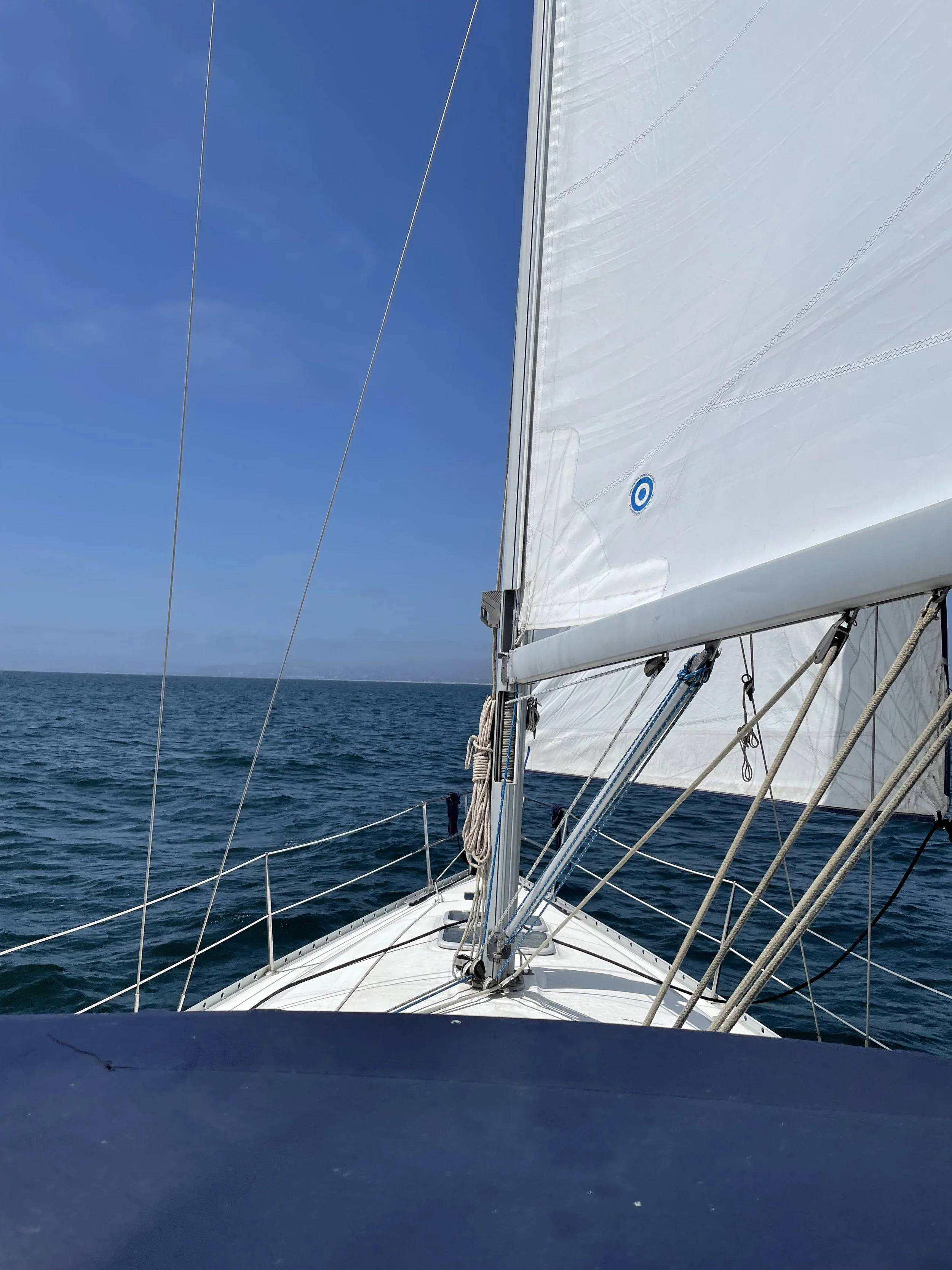 View from a sailboat at sea showing the boat's white sail, rigging, and a clear blue sky with some clouds.