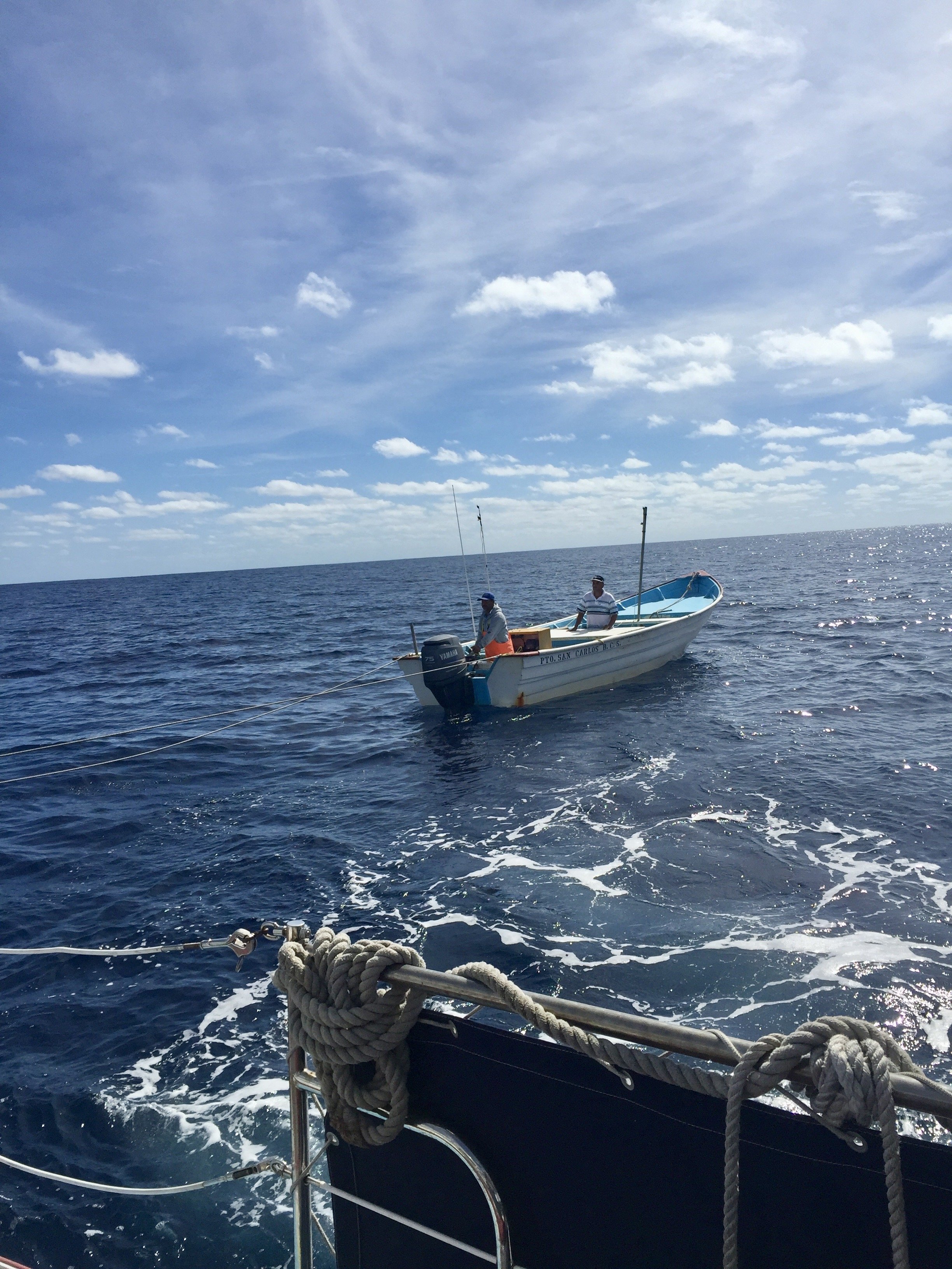 A boat on the ocean with two men, fishing rods, blue sky with scattered clouds, and a second boat in the background.