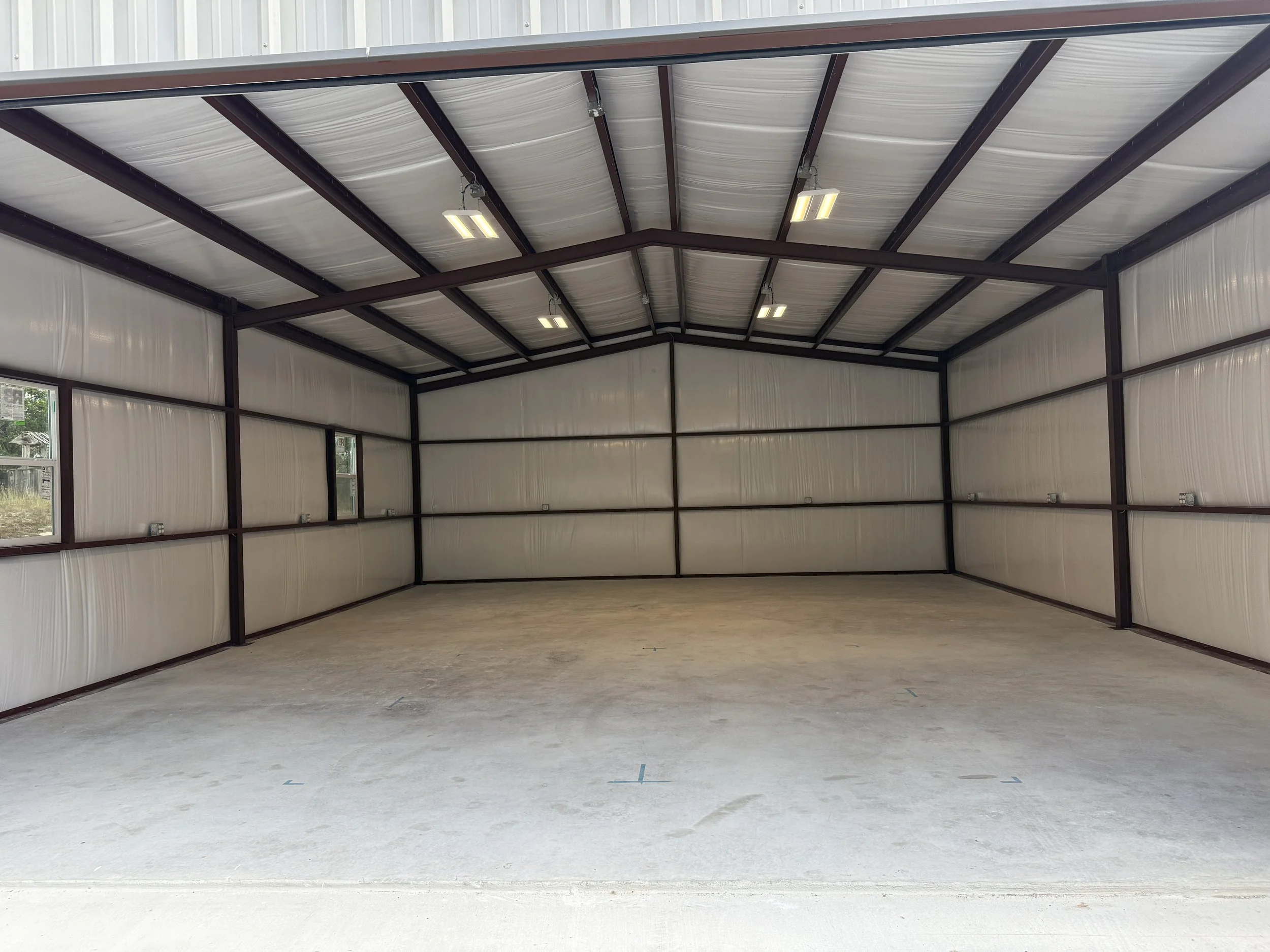 Empty interior of a metal building with a high ceiling, metal support beams, bright lighting, and a concrete floor.