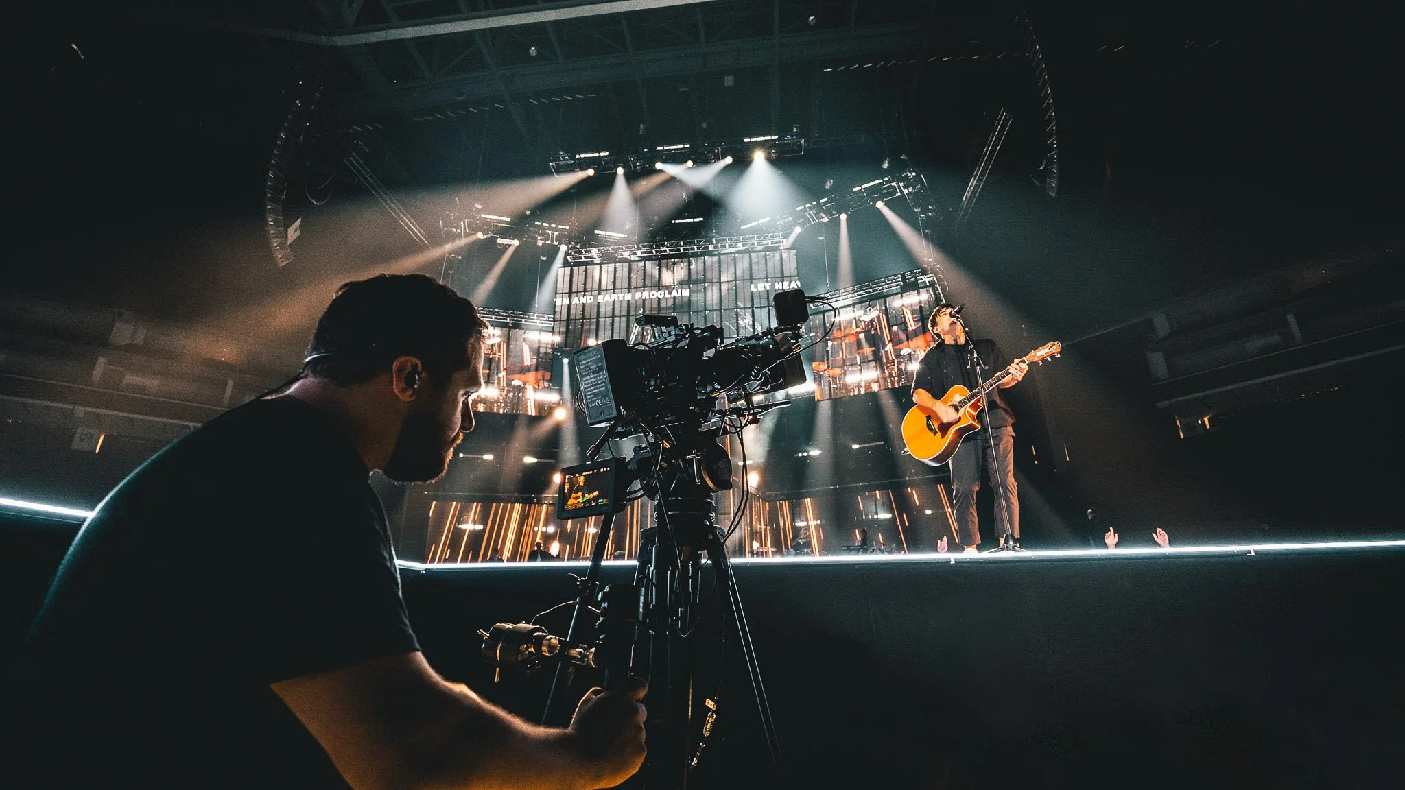 A musician playing an acoustic guitar on stage during a concert, with stage lights and digital screens in the background, and a cameraman filming the performance in the foreground.