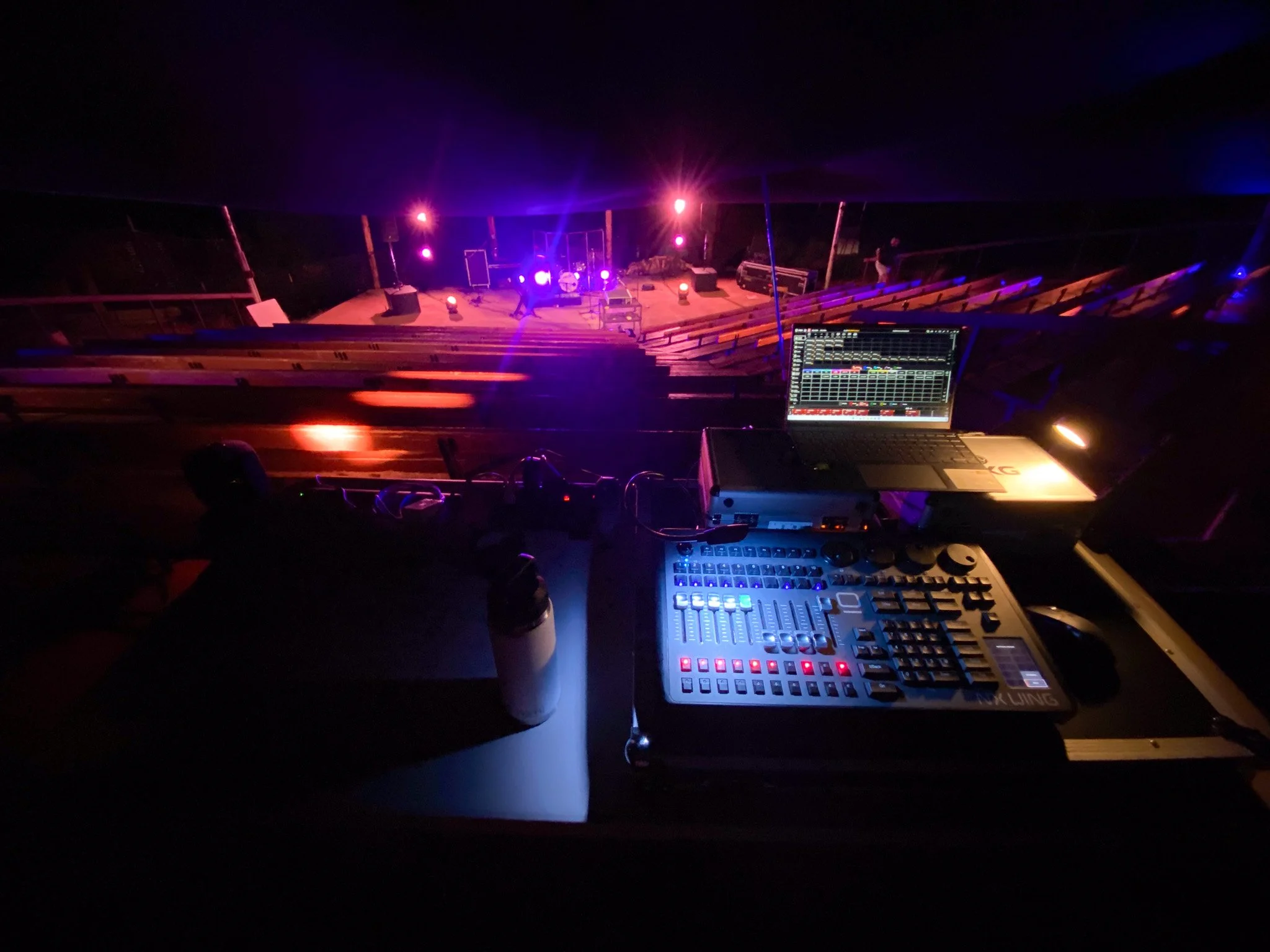 View of a stage with musical equipment, including a laptop and audio mixer, set up in a dark outdoor venue with purple and pink stage lighting.
