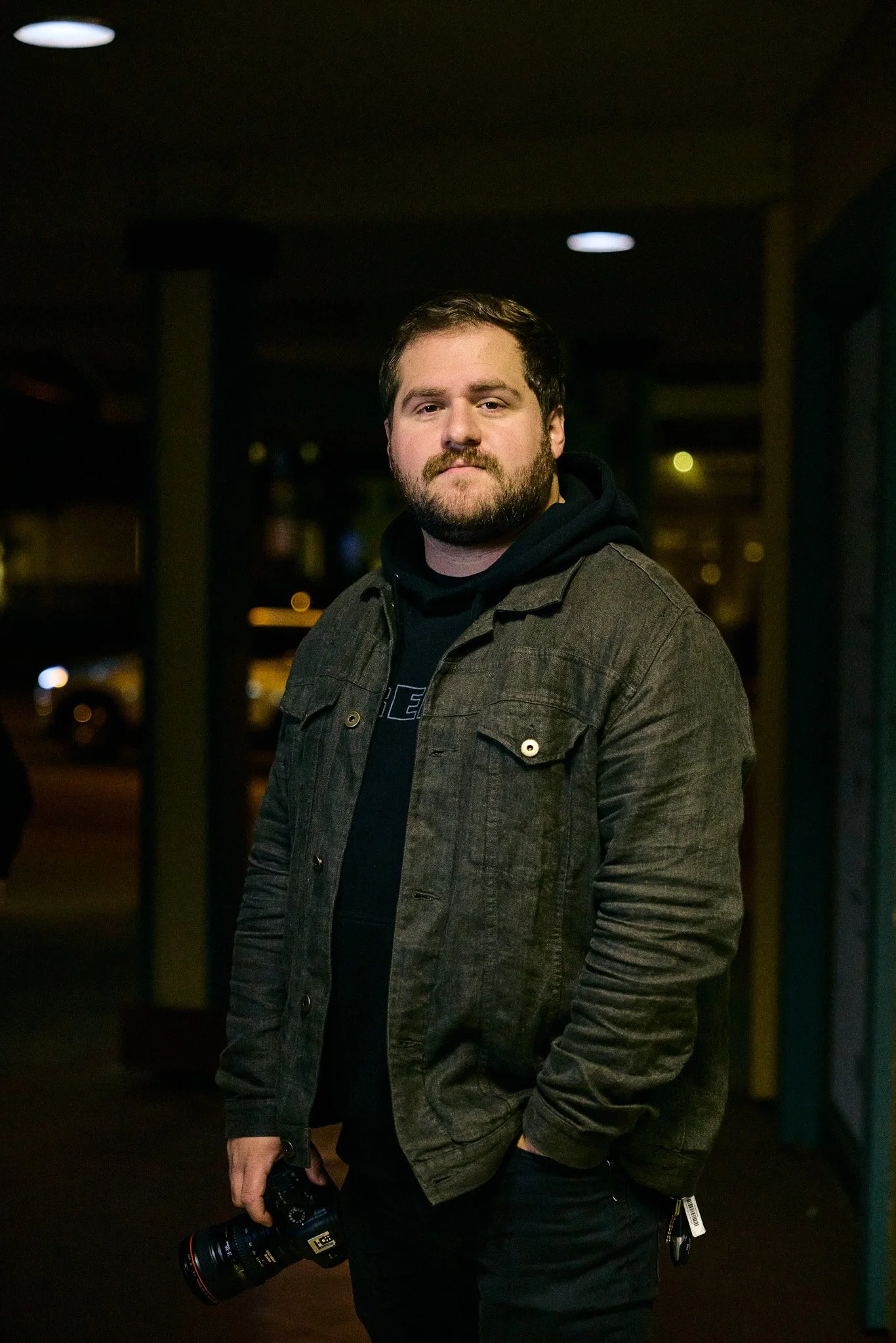 A man with a beard and mustache, wearing a black hoodie and a brown jacket, standing outside at night, holding a camera in his right hand.
