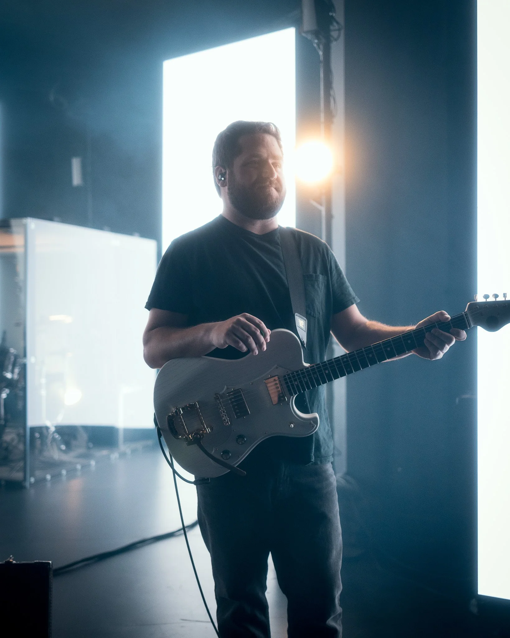 A man with a beard playing an electric guitar on stage with bright backlighting and a sunset glow behind him.