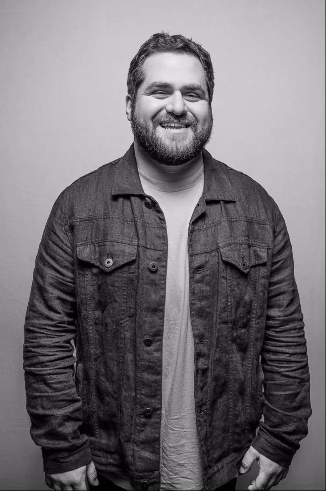 Black and white photo of a smiling man with a beard, wearing a denim jacket over a light-colored T-shirt, standing against a plain background.