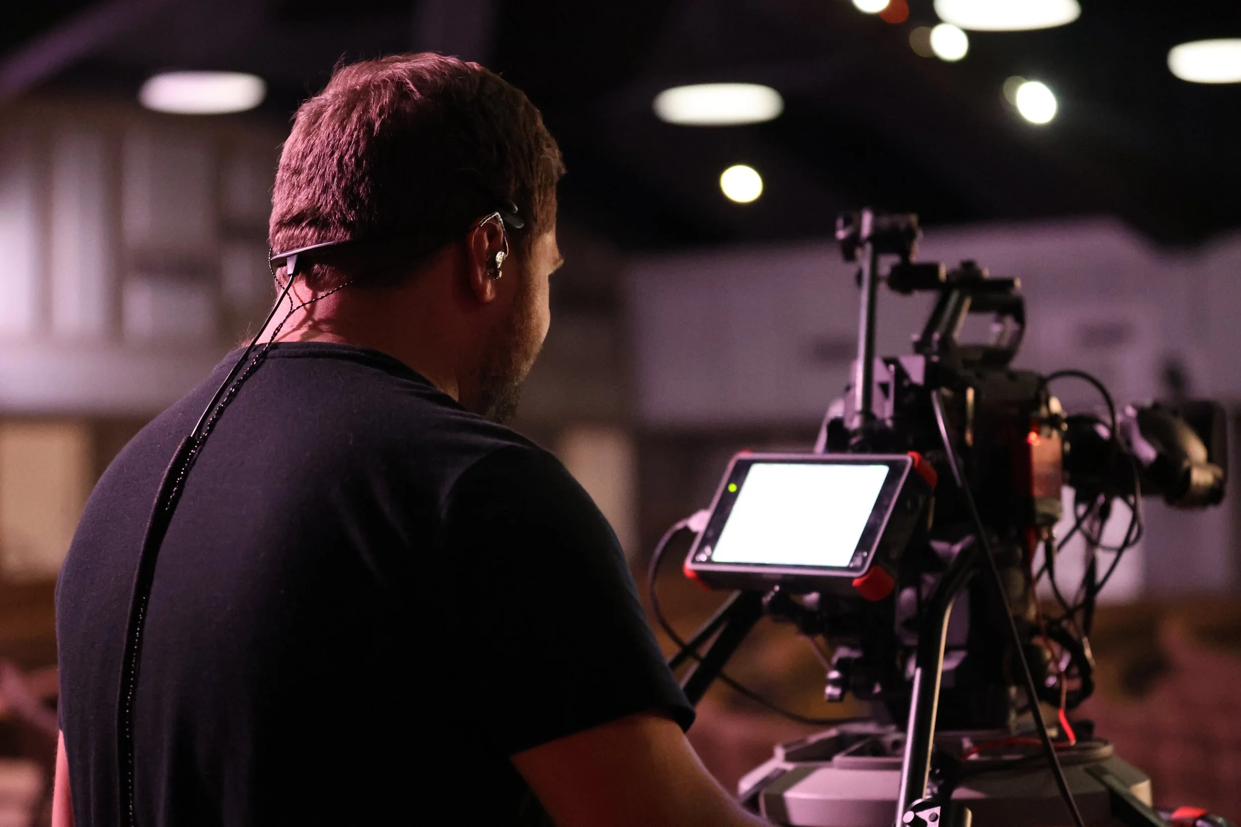 Person operating a professional camera on a tripod, with a monitor, in a dimly lit indoor setting.